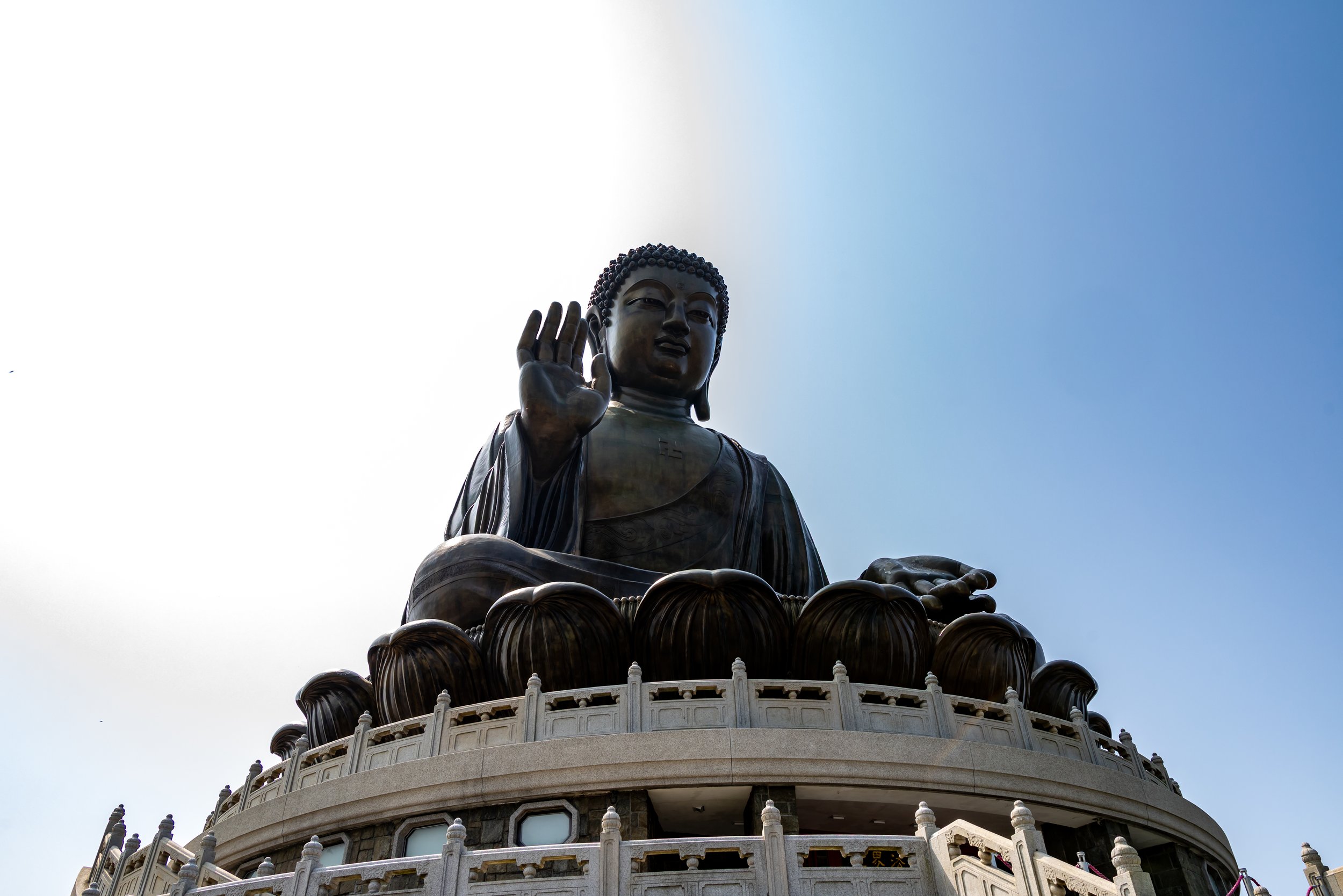 Large bronze Buddha statue seated on a decorated platform with a hand raised in a blessing gesture and the other resting on the lap, against a clear blue sky.