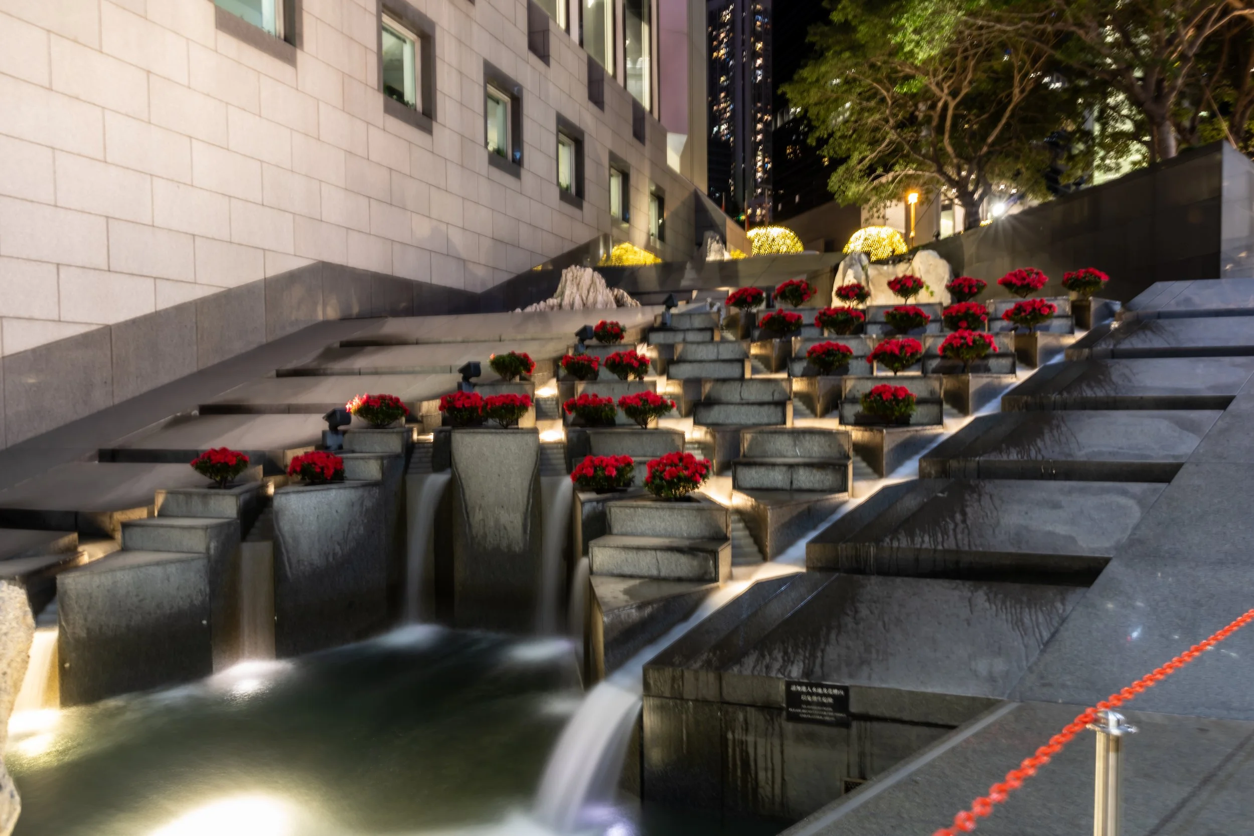 Night view of a decorative urban water feature with waterfalls and red flowering plants in tiered gray stone planters, surrounded by buildings and trees.