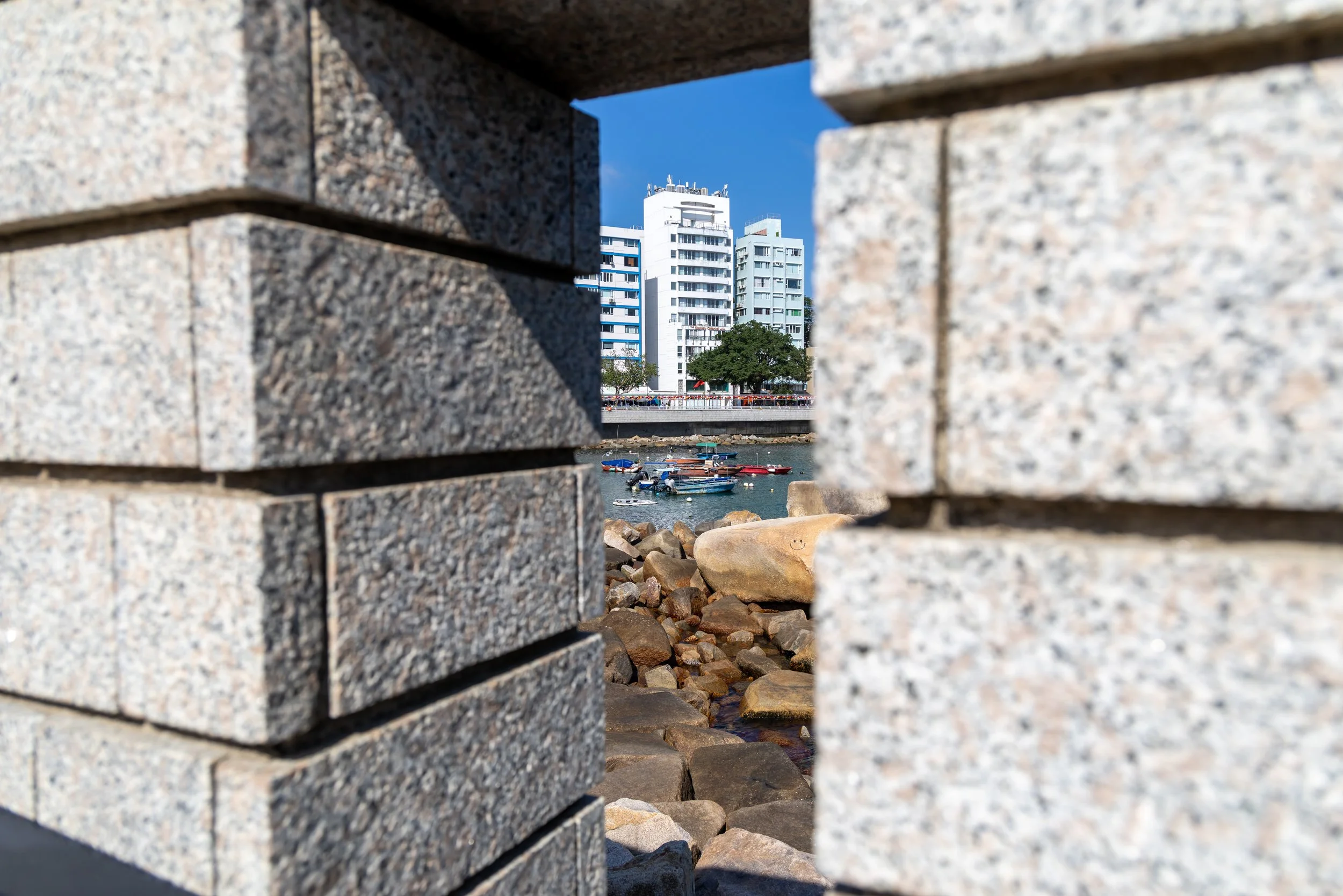 View of a cityscape with white high-rise buildings, a tree, and boats on the water, framed by brick wall openings.