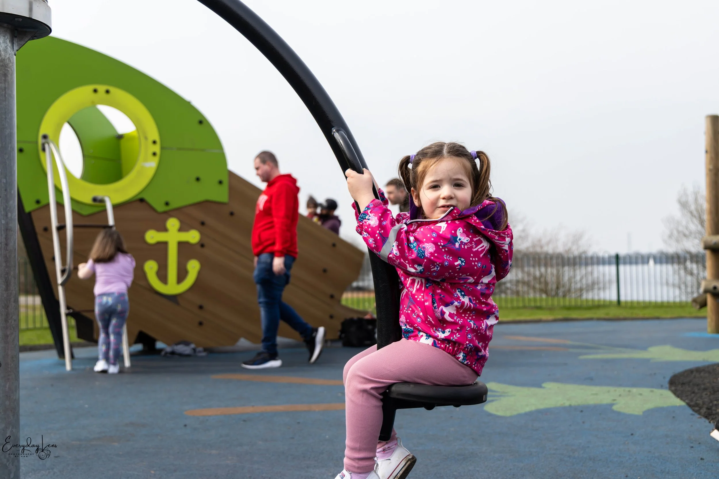 A young girl in a pink jacket sitting on a swing at a playground with a pirate ship as the background. Other children and adults are seen in the background near the pirate ship.
