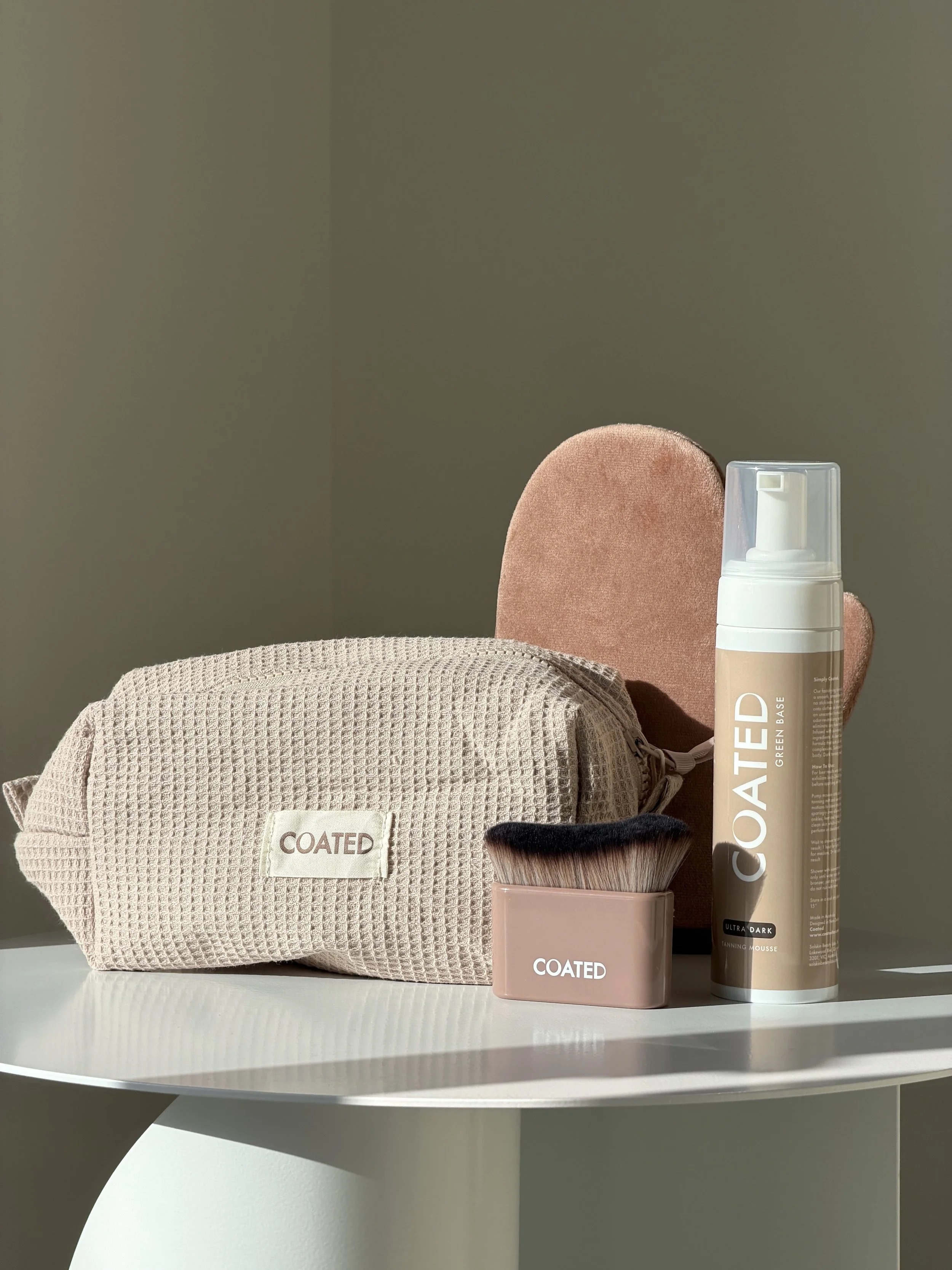Beauty products including a waffle-textured beige bag, a small bag with a makeup brush labeled 'COATED', and a large bottle of 'COATED' green base tan, on a white table with a tan mitt in the background.