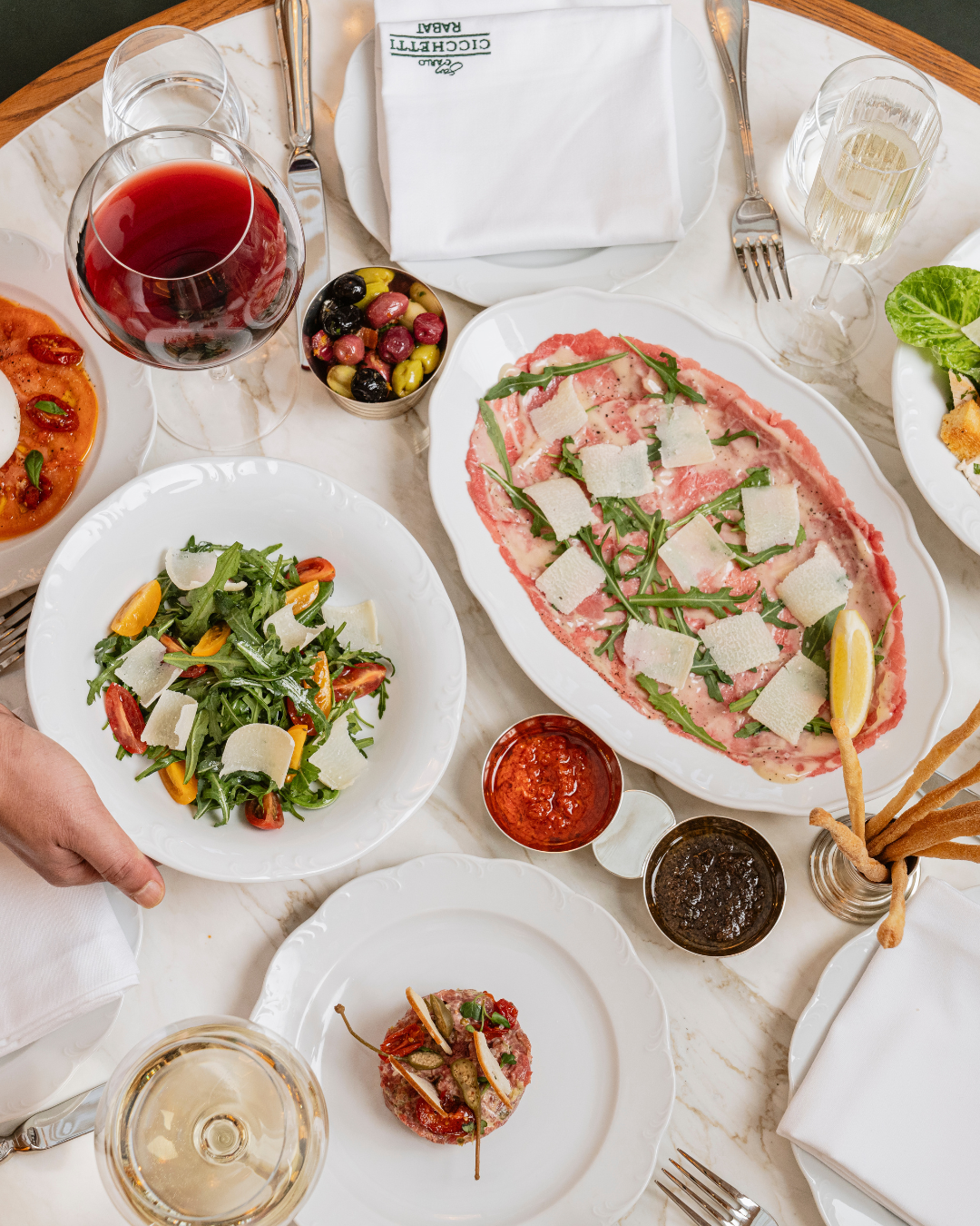 Repas de restaurant avec assiettes de carpaccio, salade, olives, sauces, verre de vin blanc et verre de vin rouge sur une table en marbre blanc.