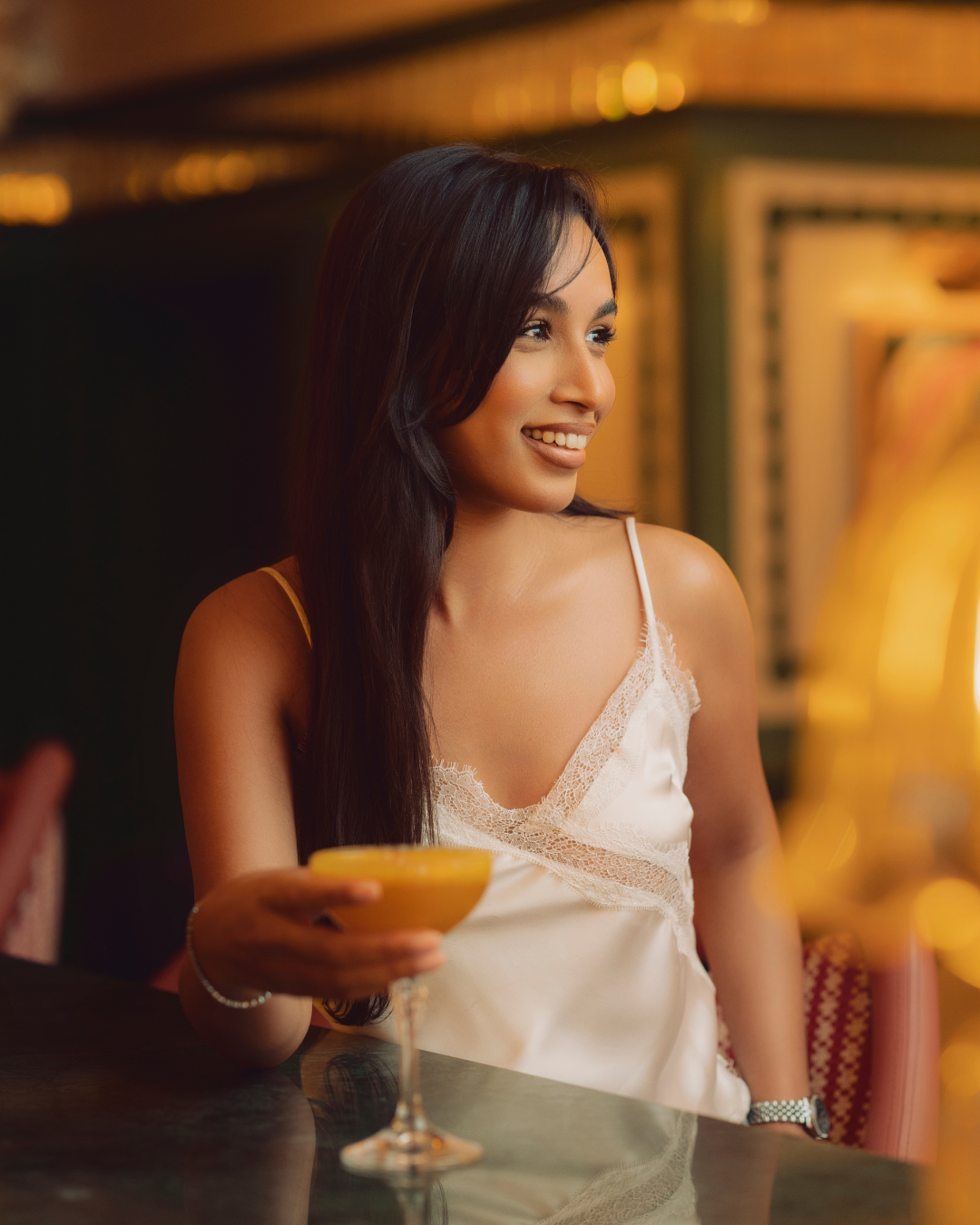 Une femme souriante avec des cheveux longs noirs, portant une robe en satin blanc, tient un cocktail dans un verre à pied, dans un décor élégant et chaleureux.
