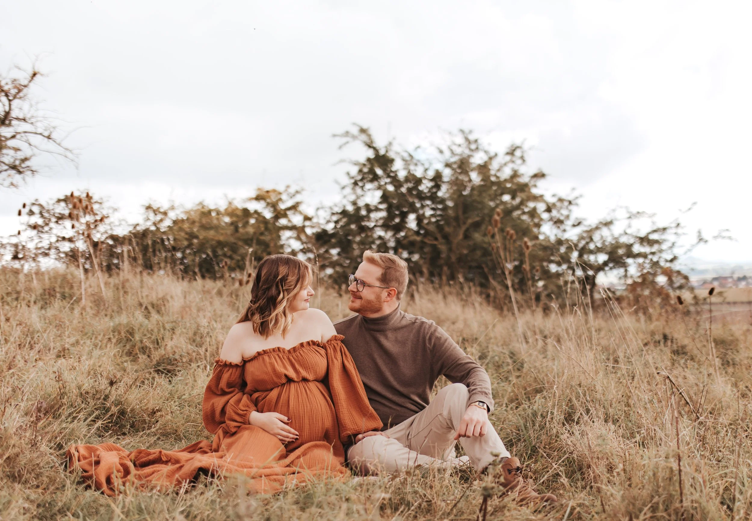 Ein schwangertes Paar sitzt im Gras in einer natürlichen Umgebung und schaut sich liebevoll an. Sie ist in einem braunen, off-shoulder Kleid, er trägt einen grauen Pullover und beige Hosen.
