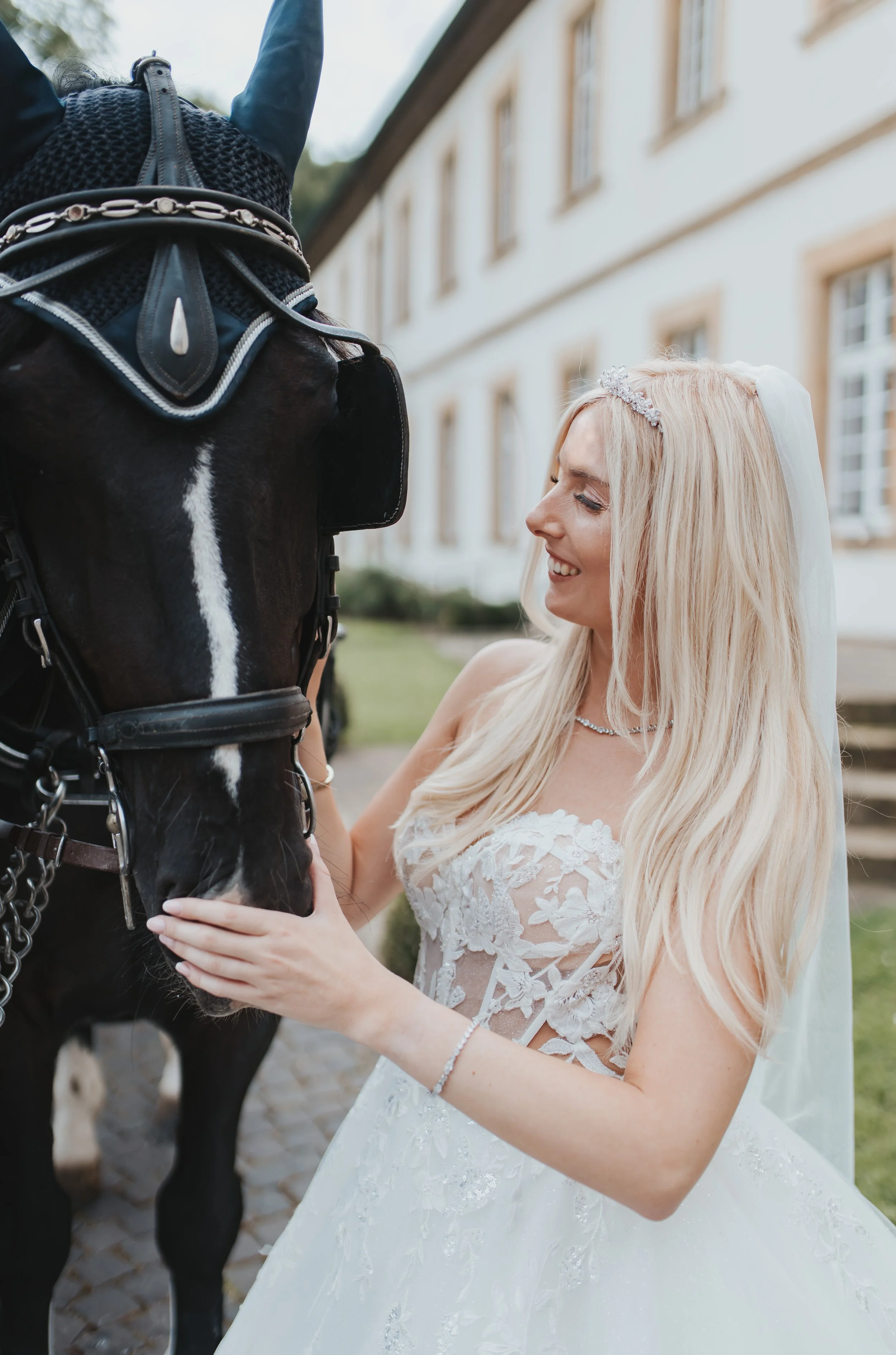 Braut in Kleid und Tiara streichelt ein schwarzes Pferd vor einem Gebäude.