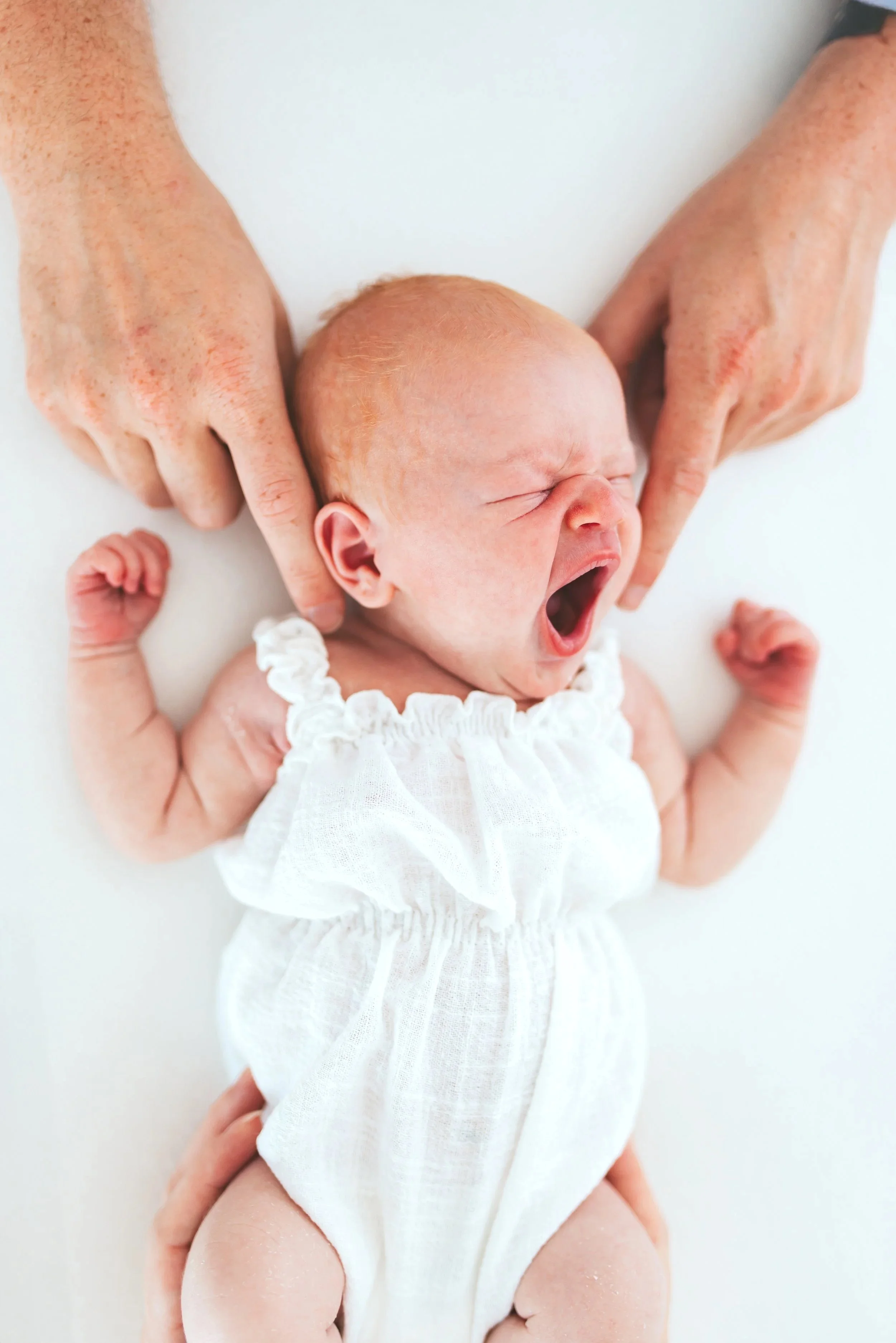 Ein schreiendes Baby in einem weißen Kleid, gehalten von zwei Erwachsenen, mit nur den Händen sichtbar.