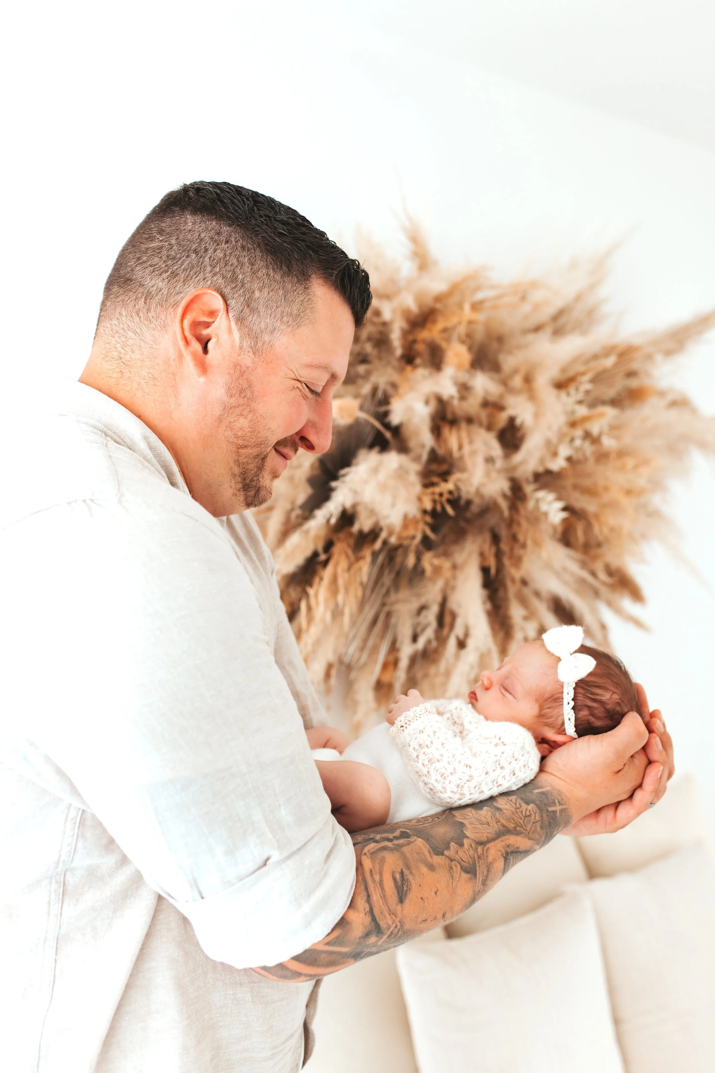 Ein Mann hält ein neugeborenes Baby in den Händen, beide lachen und schauen sich an. Im Hintergrund sind getrocknetes Pampasgras.