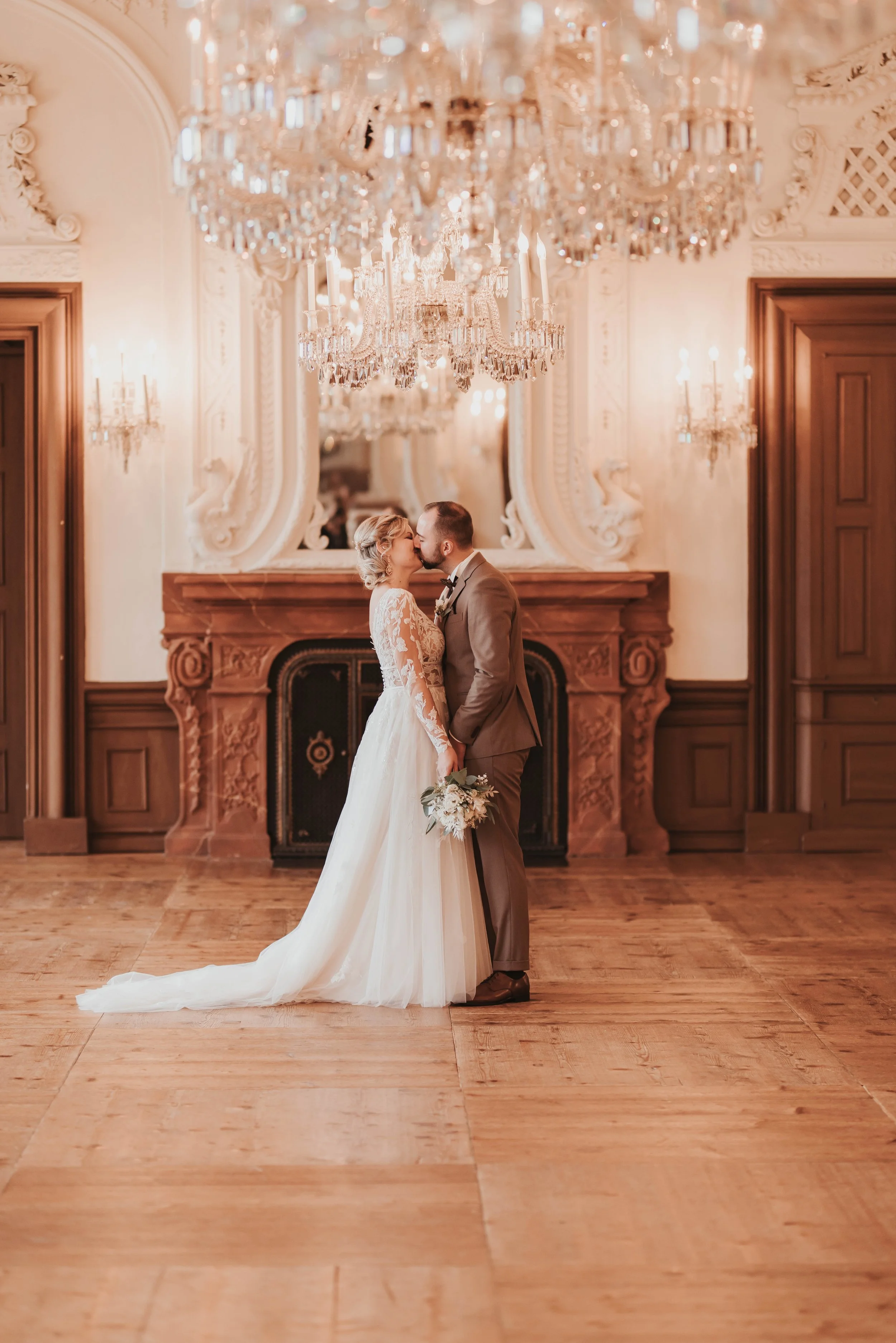 Braut und Bräutigam küssen sich in einem eleganten Saal mit Kronleuchtern und Holzverkleidungen.