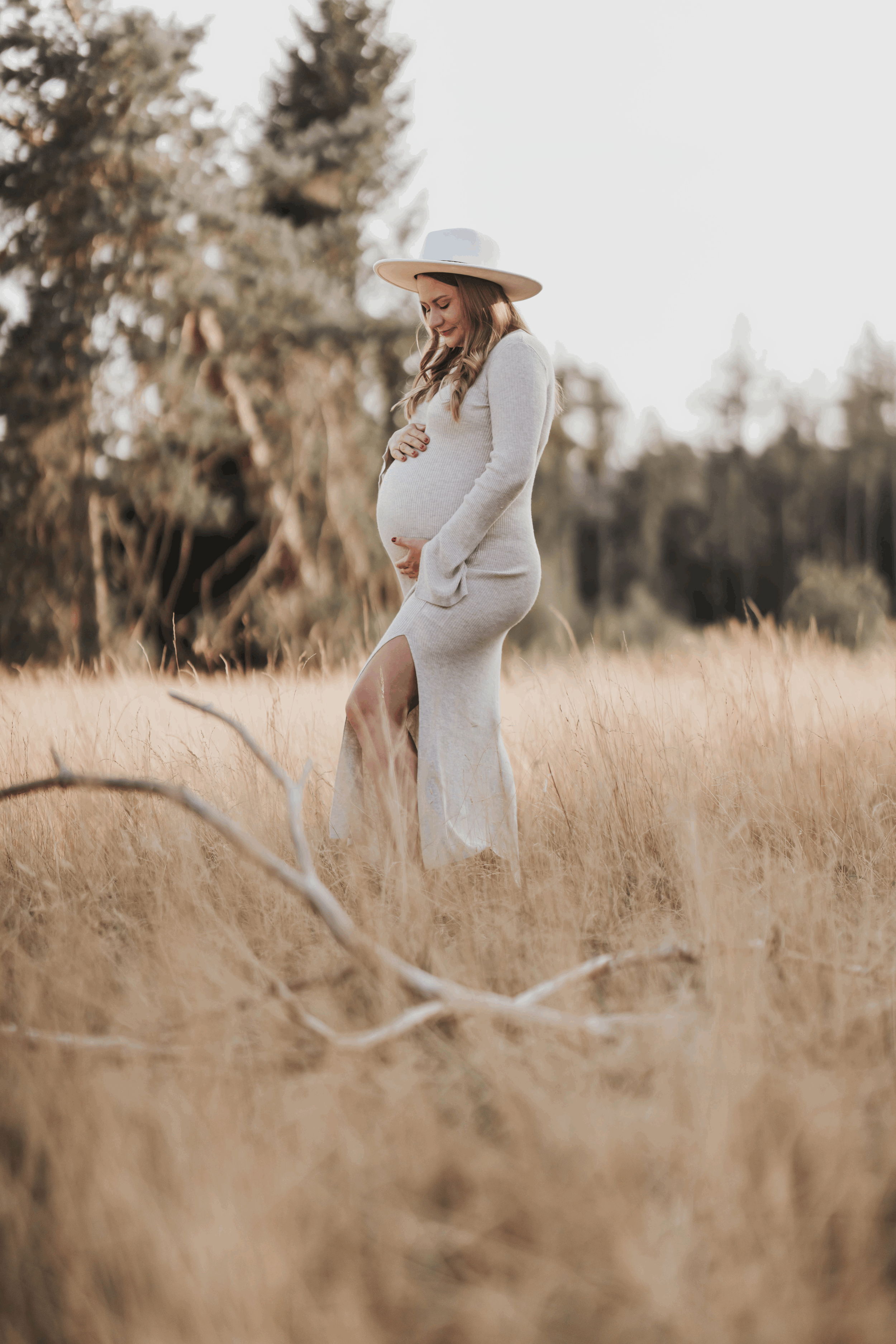 Schwangere Frau in langem grauem Kleid und Hut in einer goldenen Graslandschaft, die Hand auf den Bauch legt und lächelt.