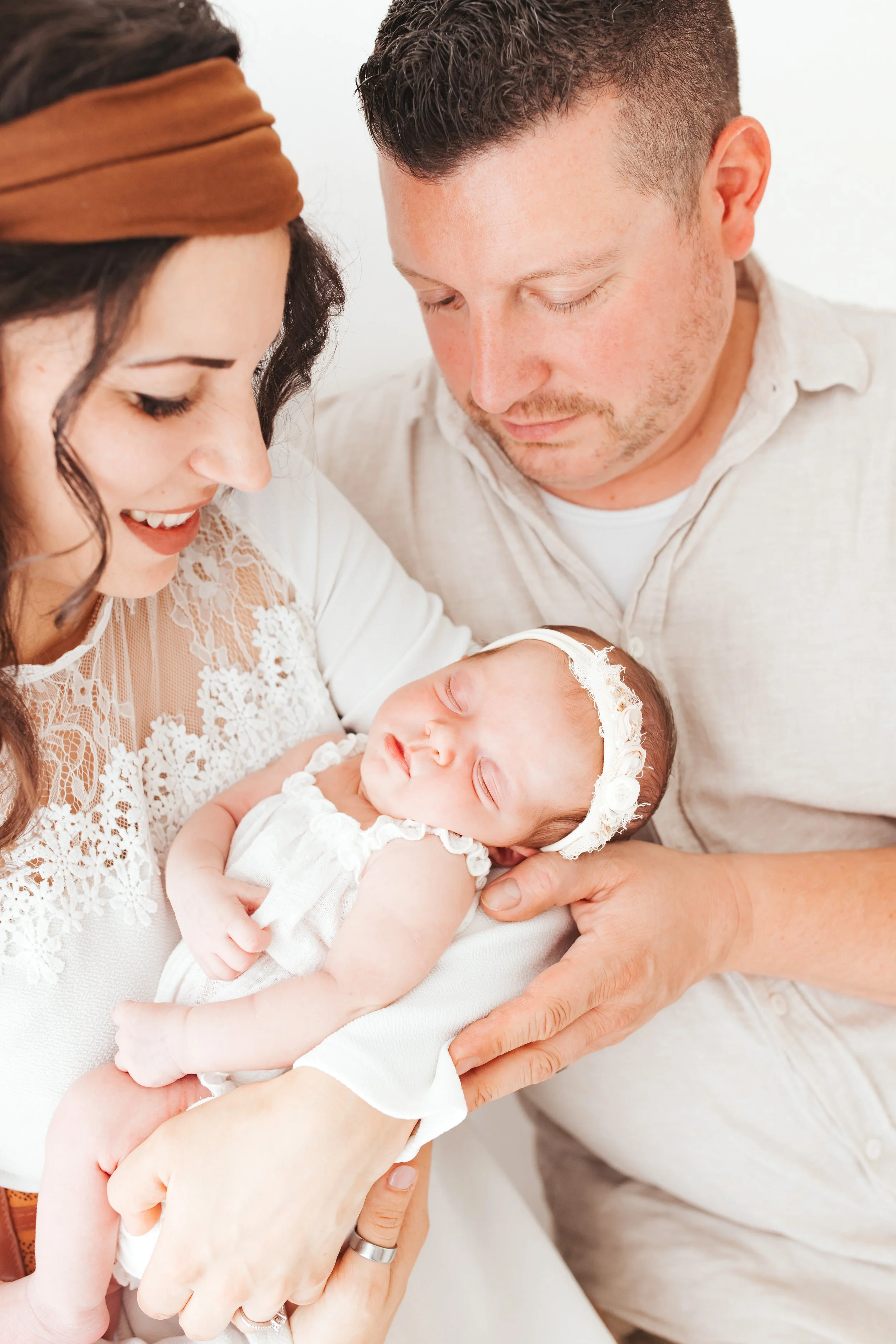 Ein Baby wird von einer glücklichen Familie gehalten, bestehend aus einer Frau, einem Mann und dem Baby, alle in hellen, weißen Kleidung. Das Baby trägt eine weiße Haarkrone und ist schlafend.