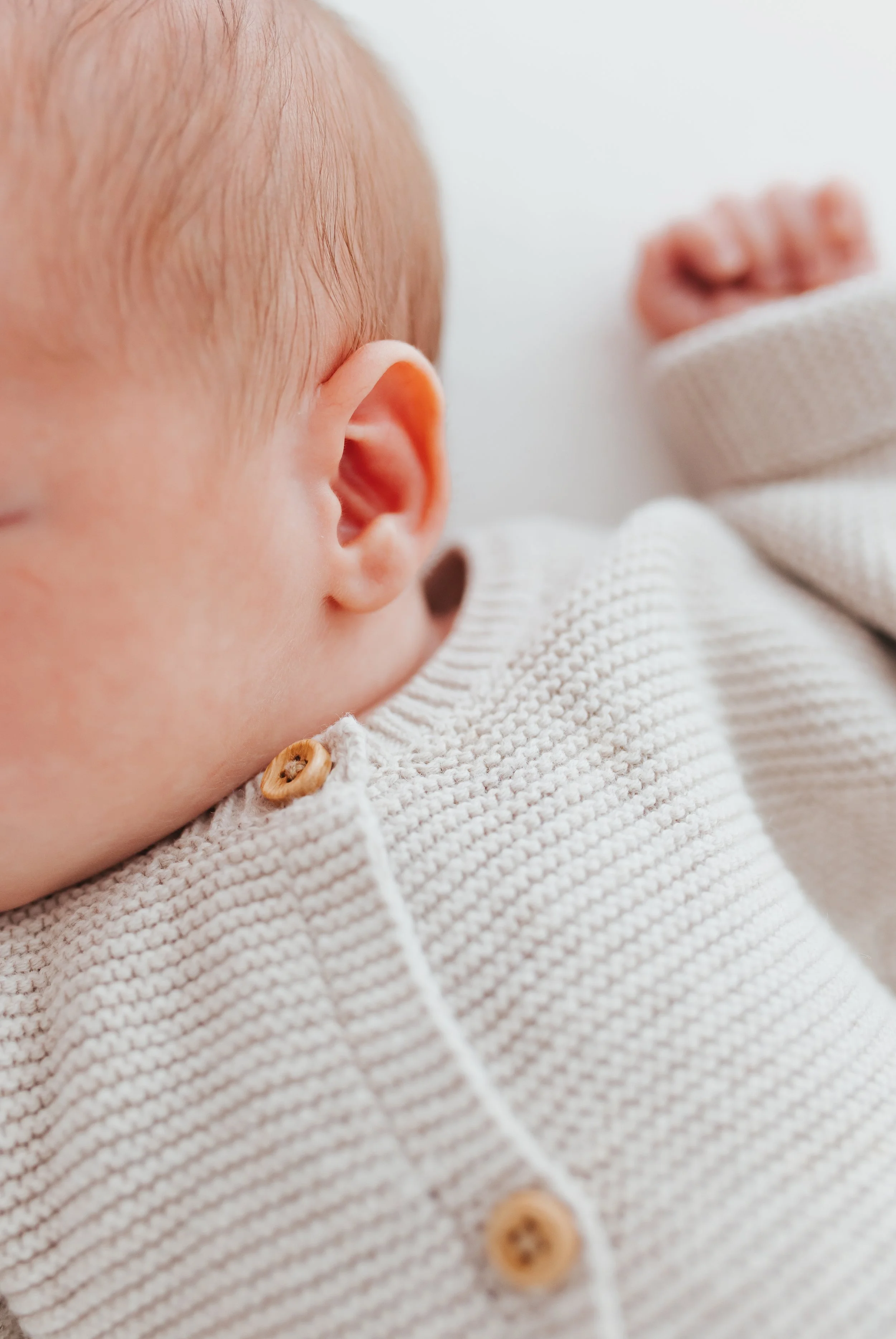 Nahaufnahme eines Babys, das auf seiner Seite liegt, mit Blick auf sein Ohr und Teil seines Gesichts, das in einem weißen, gestrickten Pullover steckt.