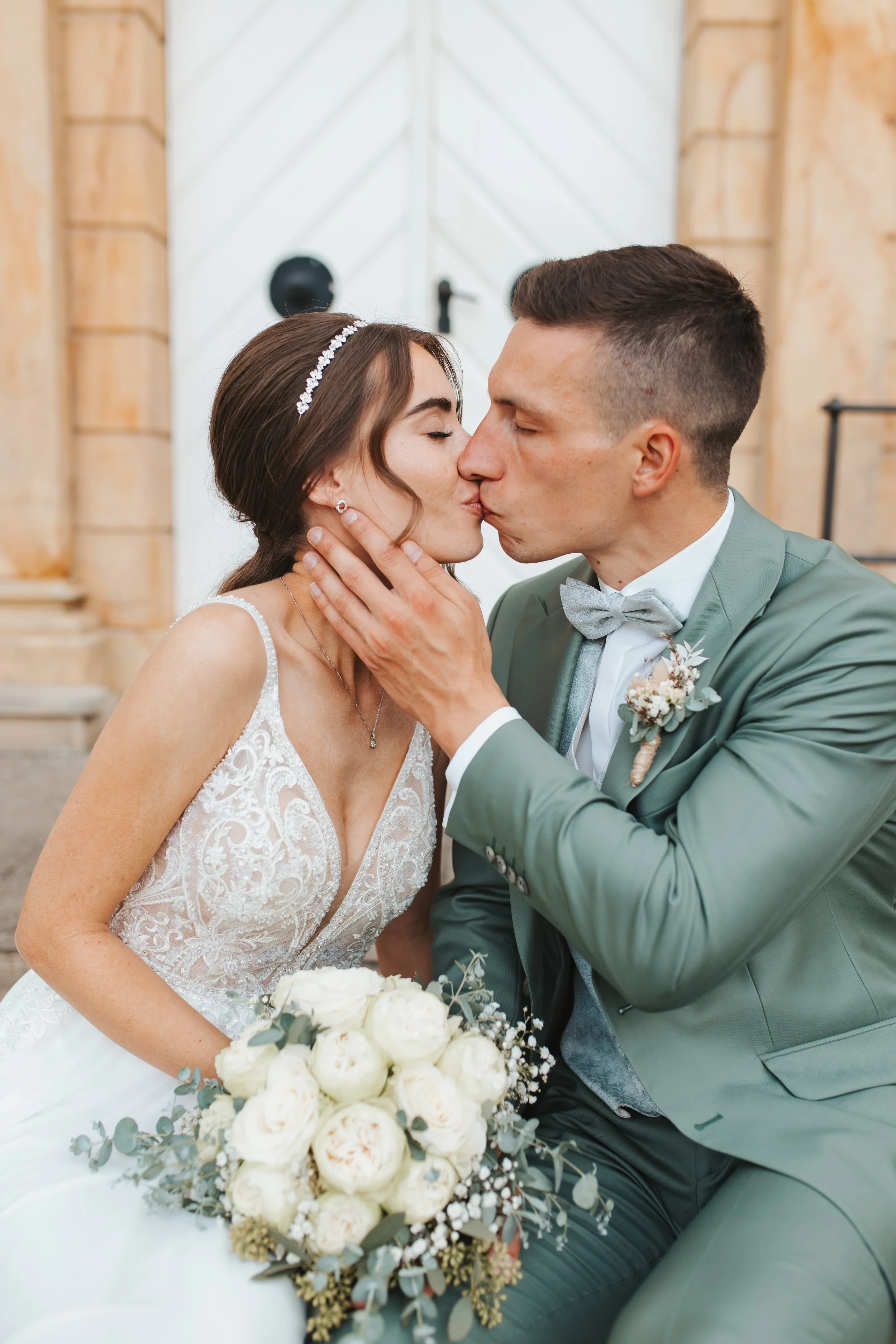 Ein Brautpaar bei einer Hochzeitszeremonie, das sich küsst, der Brautstrauß in ihrer Hand und die Braut in einem weißen Kleid mit Spitze, der Bräutigam in einem grünen Anzug mit Fliege.