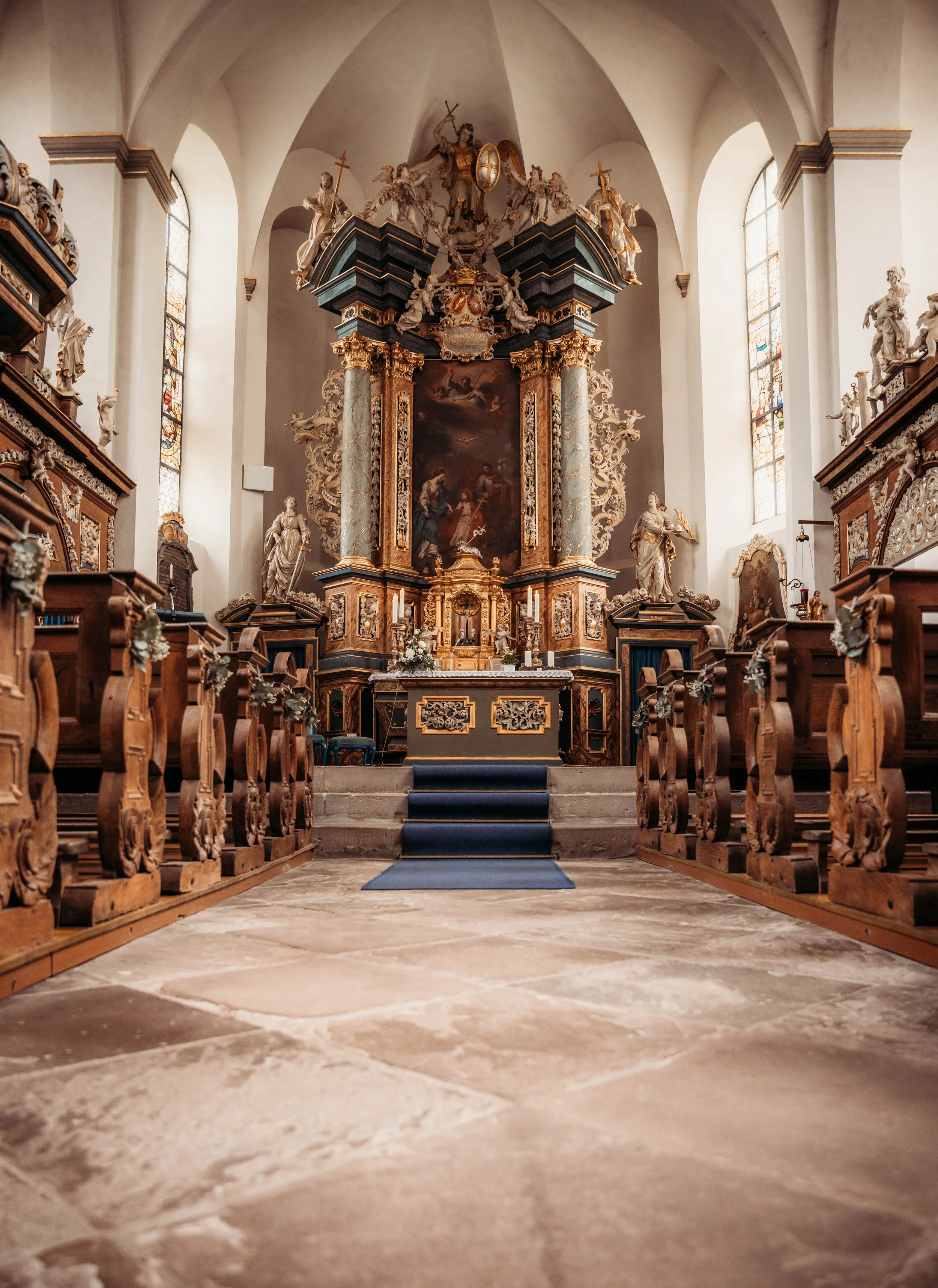 Innenraum einer Barockkirche mit Altar im Hintergrund, barocke Gegenstände, Skulpturen, rotgoldene Verzierungen, große Fenster, Holzsitze auf beiden Seiten, blauer Teppich auf den Stufen zum Altar.