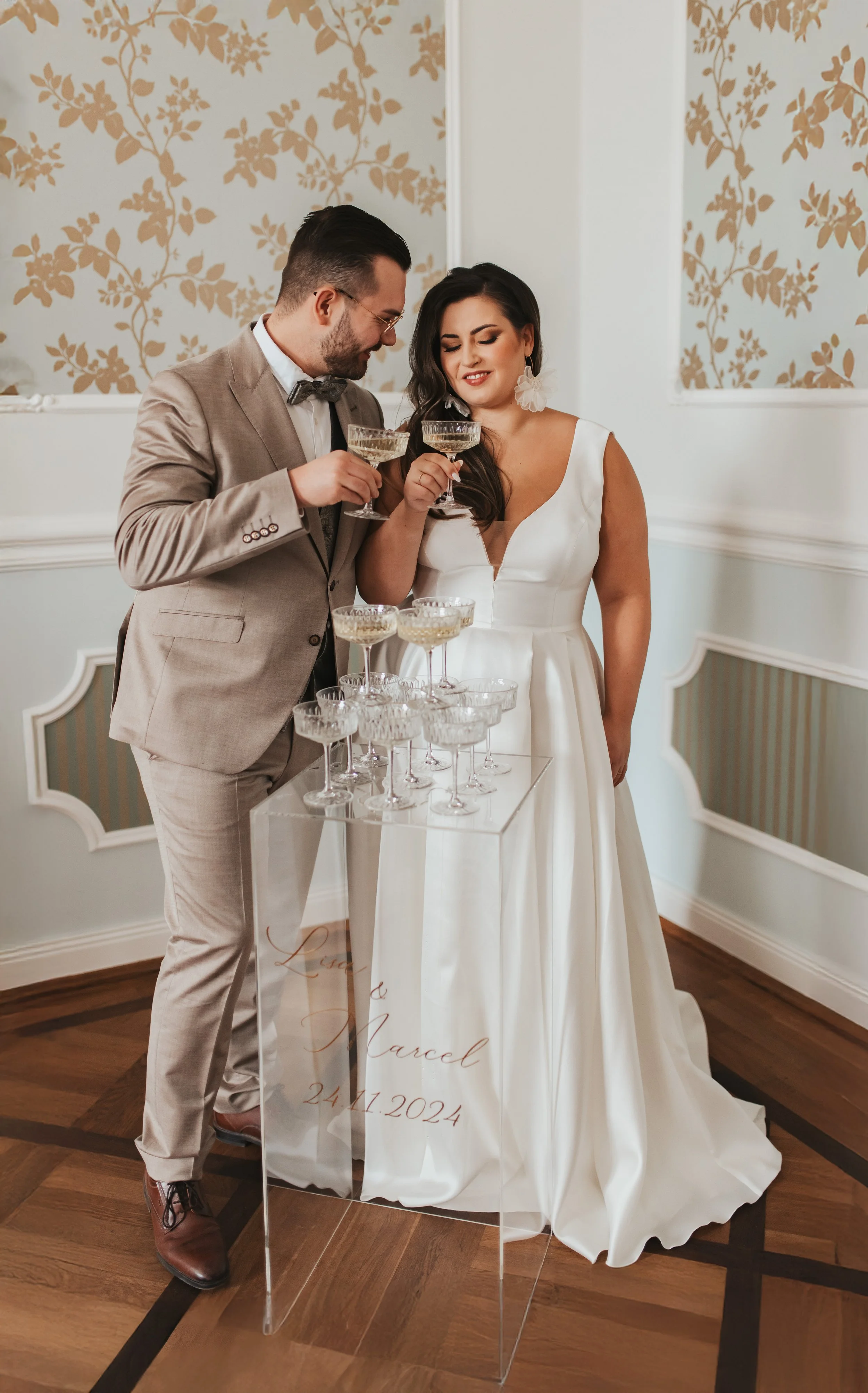 Ein Brautpaar beim Anstoßen mit Champagnergläsern, stehend hinter einem transparenten Tisch mit mehreren Champagnergläsern, bei einer Hochzeit am 24.11.2024.