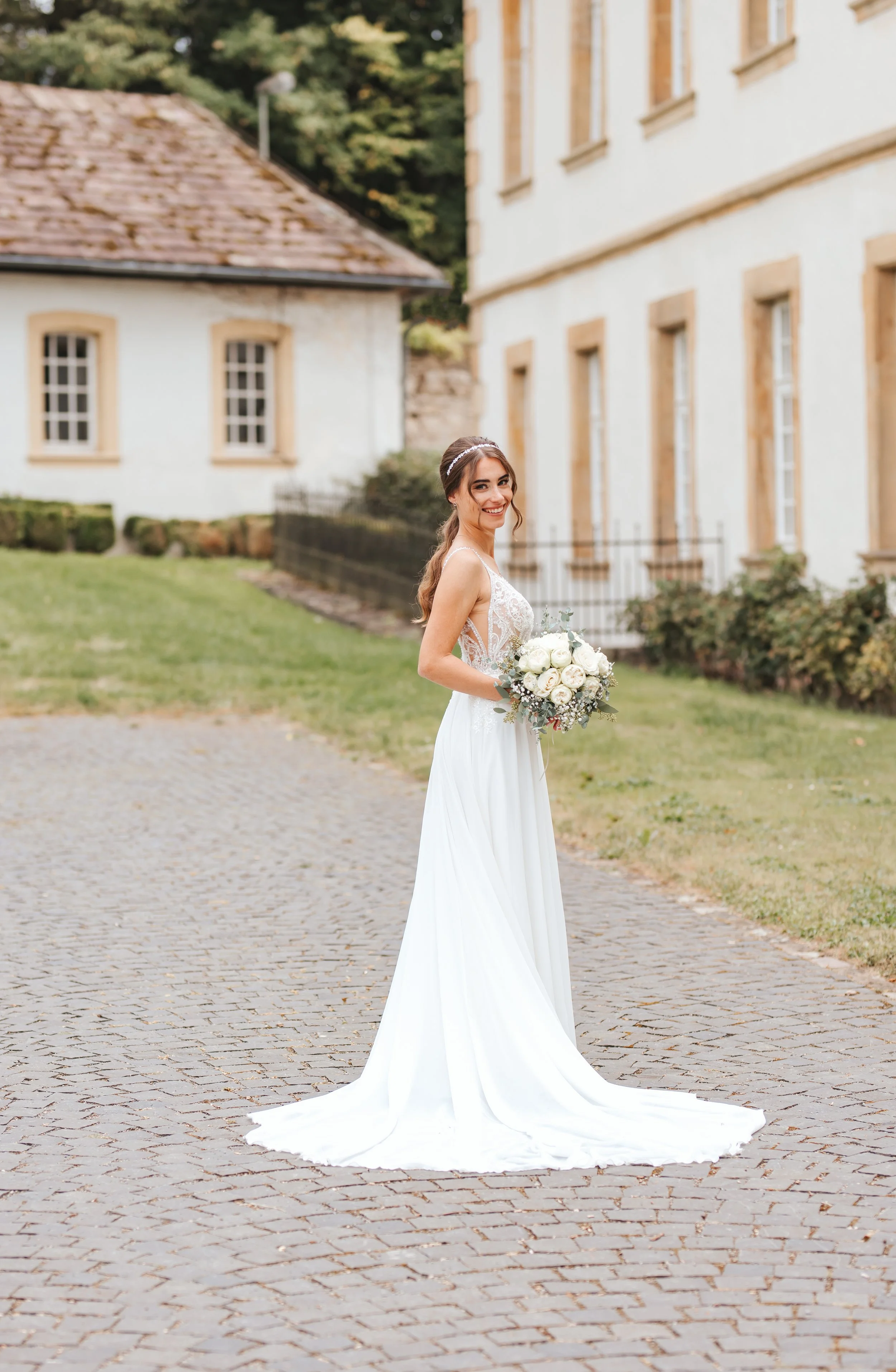 Braut in weißem Hochzeitskleid hält einen Blumenstrauß und steht auf einer gepflasterten Straße vor historischen Gebäuden, im Freien bei Tageslicht.