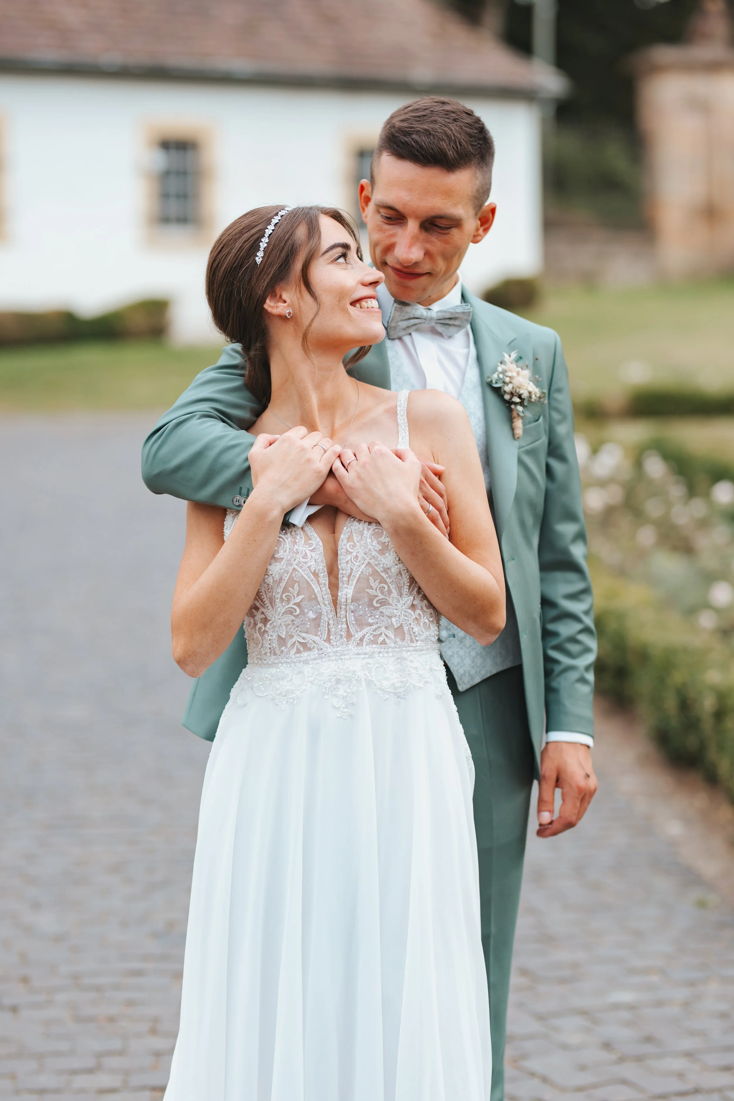 Ein frisch verheiratetes Paar, die Braut in einem weißen Hochzeitskleid mit Spitze, der Bräutigam in einem grünen Anzug, umarmt sich vor einem Haus im Freien.
