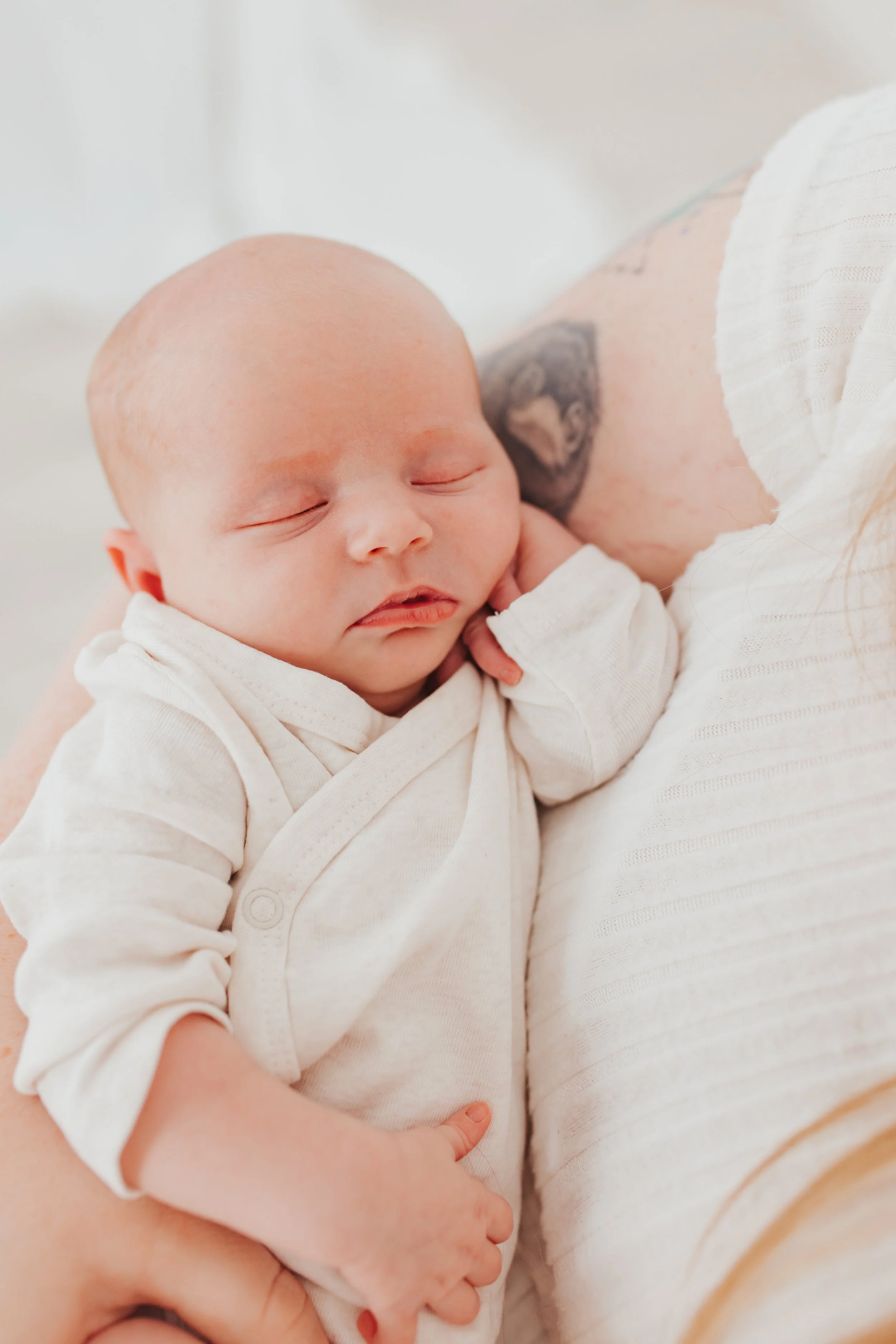Ein schlafender Baby in weißen Kleid auf einer Person in einem hellen Raum.