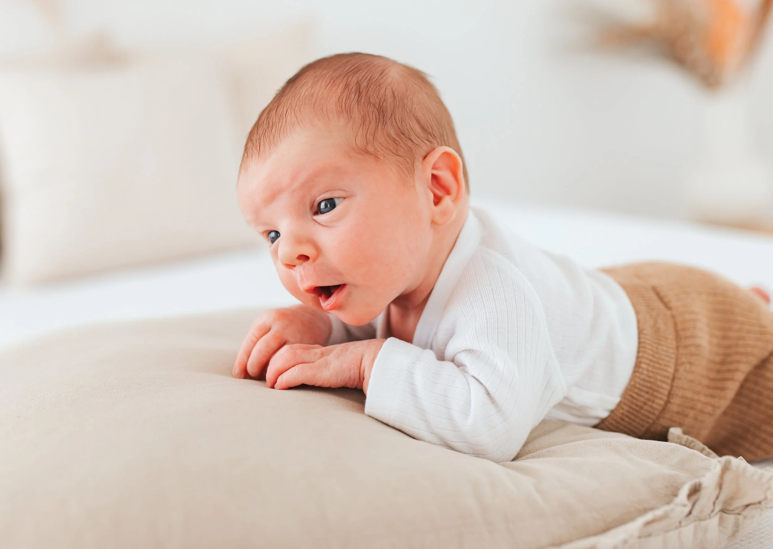 Ein Baby liegt auf dem Bauch auf einem weichen Kissen, schaut neugierig nach links, trägt ein weißes Oberteil und bequeme braune Hosen, in einem hellen Raum.