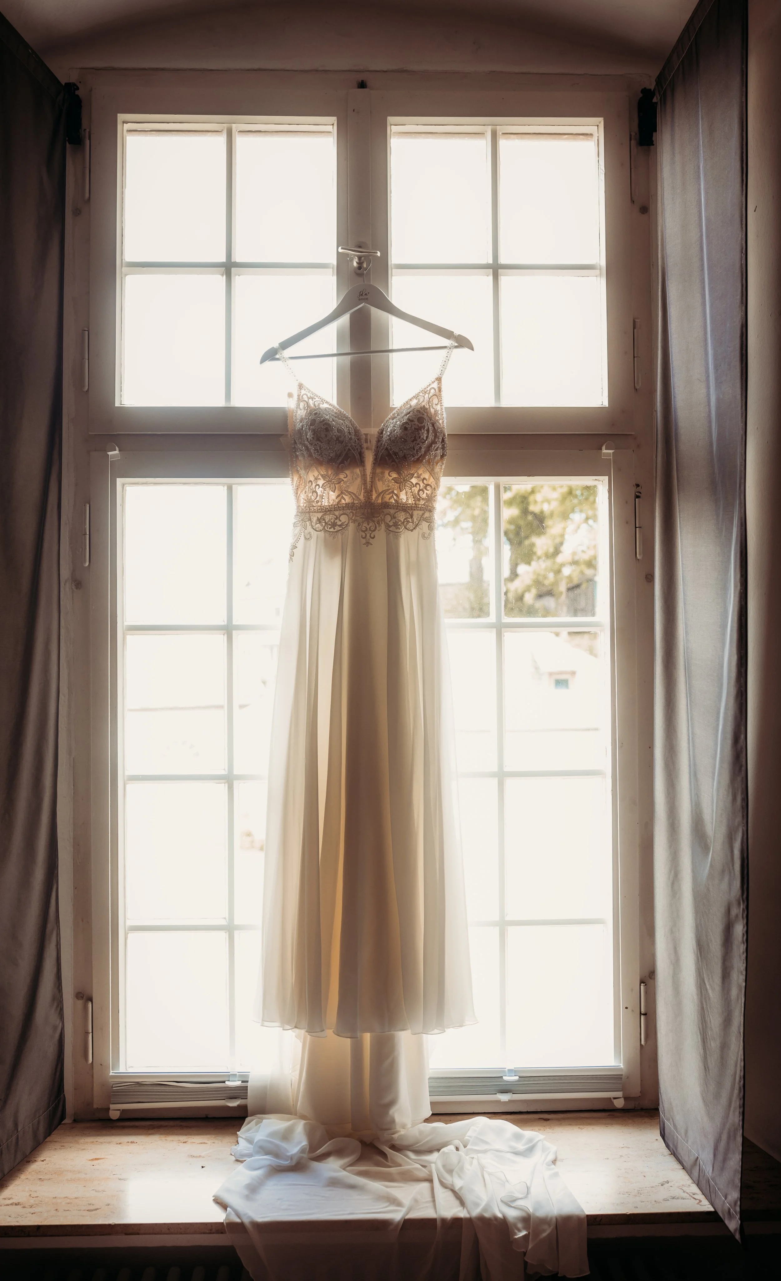 Ein Brautkleid hängt an einem Bügel vor einem hellen Fenster