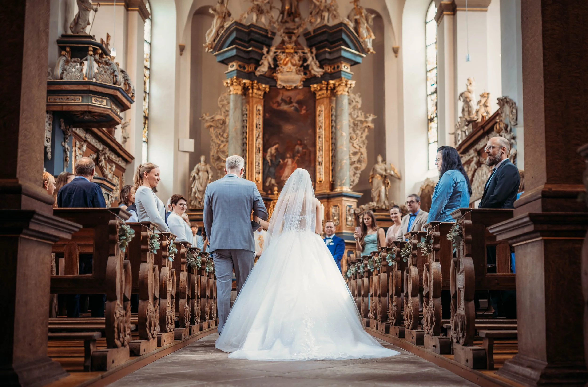 Braut und Bräutigam bei der Hochzeit in einer barocken Kirche, umgeben von Gästen.