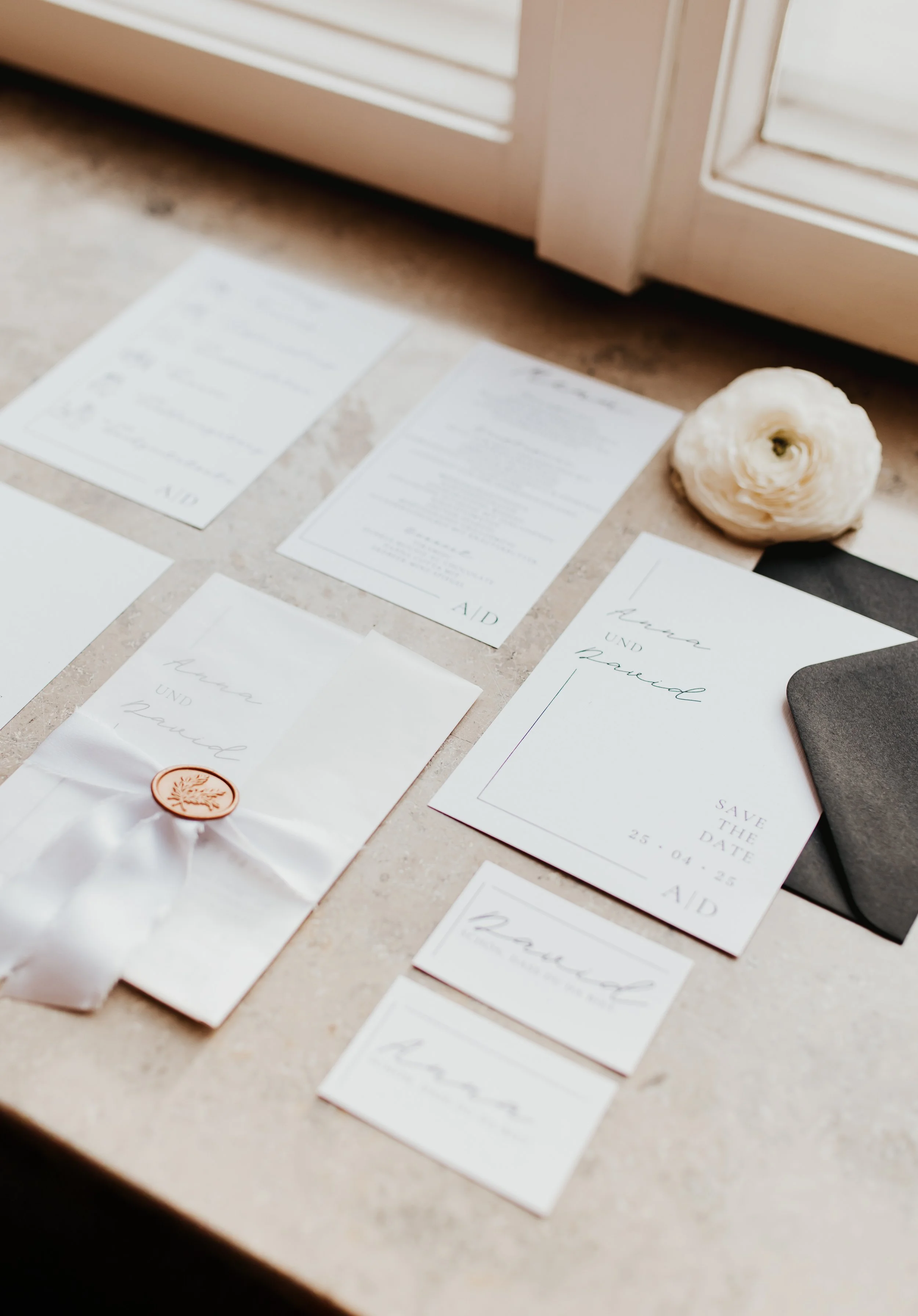 Verschiedene Hochzeitskarten, ein Briefumschlag mit Siegel, eine weiße Blüte und ein schwarzer Umschlag liegen auf einer Steinoberfläche.