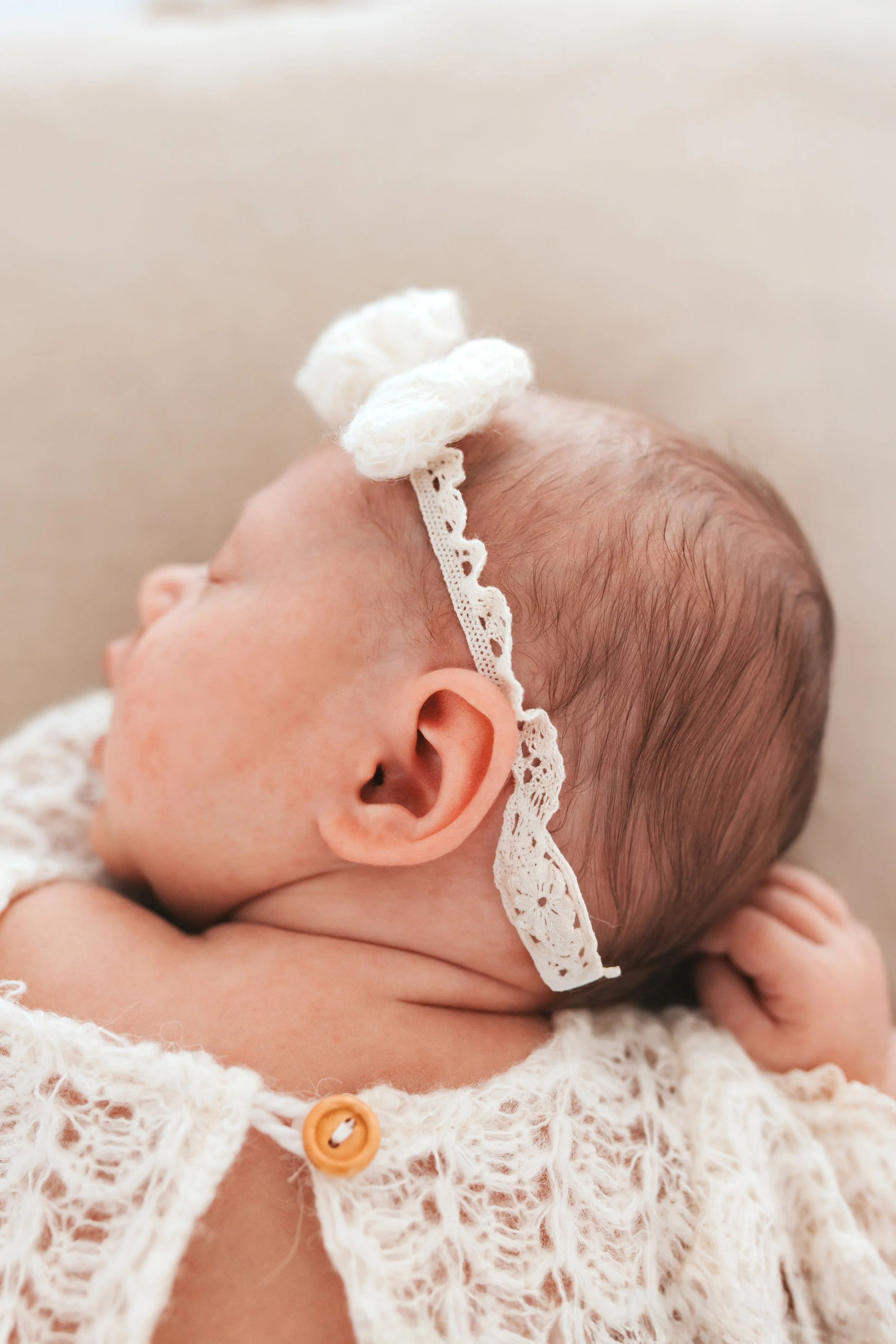 Ein neugeborenes Baby liegt auf der Seite, trägt eine weiße, handgestrickte Kleidung und eine weiße, gehäkelte Haarschleife.