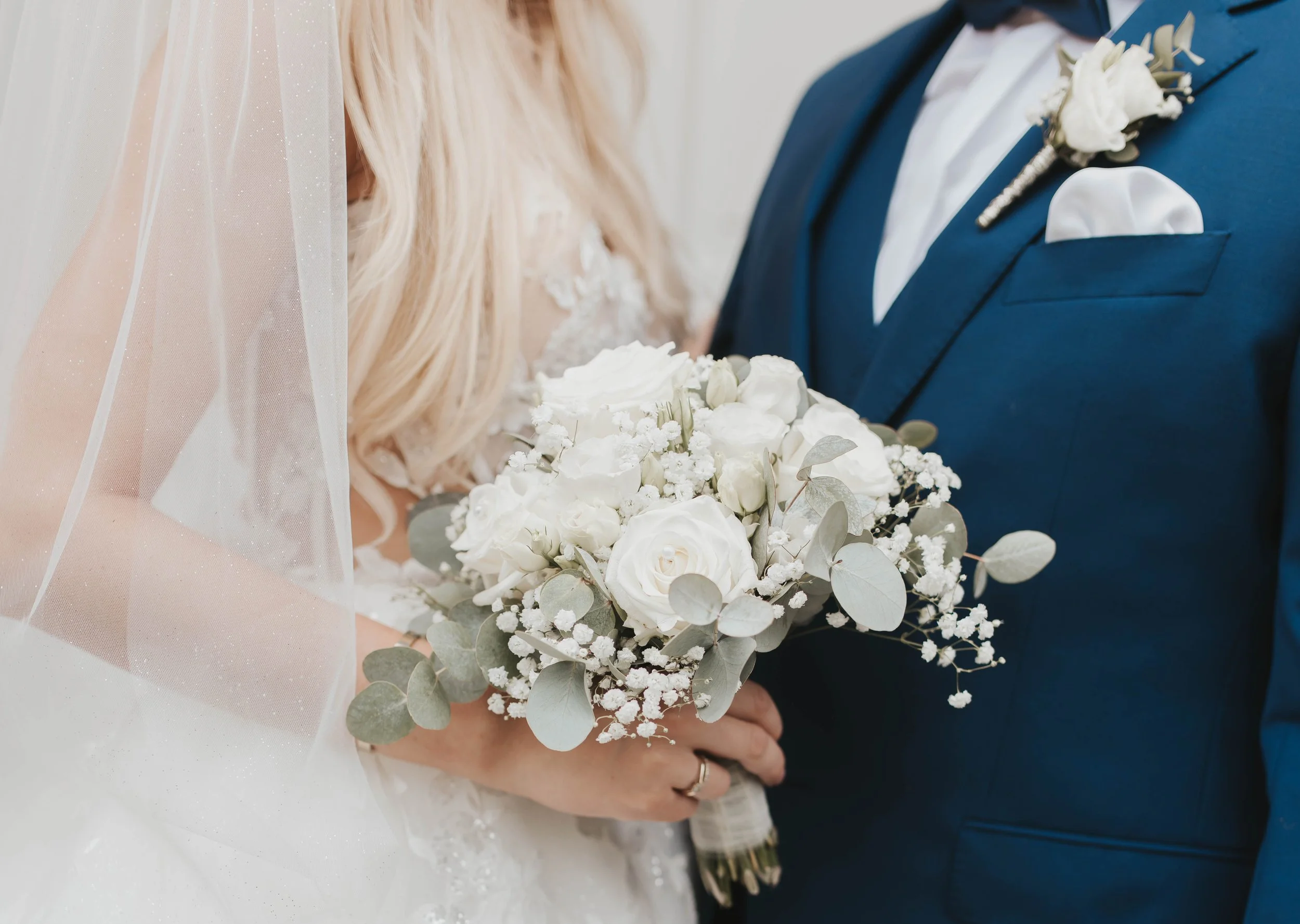 Nahaufnahme eines Brautpaares bei einer Hochzeit, die Braut hält einen weißen Blumenstrauß, der Bräutigam trägt einen blauen Anzug mit weißem Hemd und einer weißen Blume im Revers.