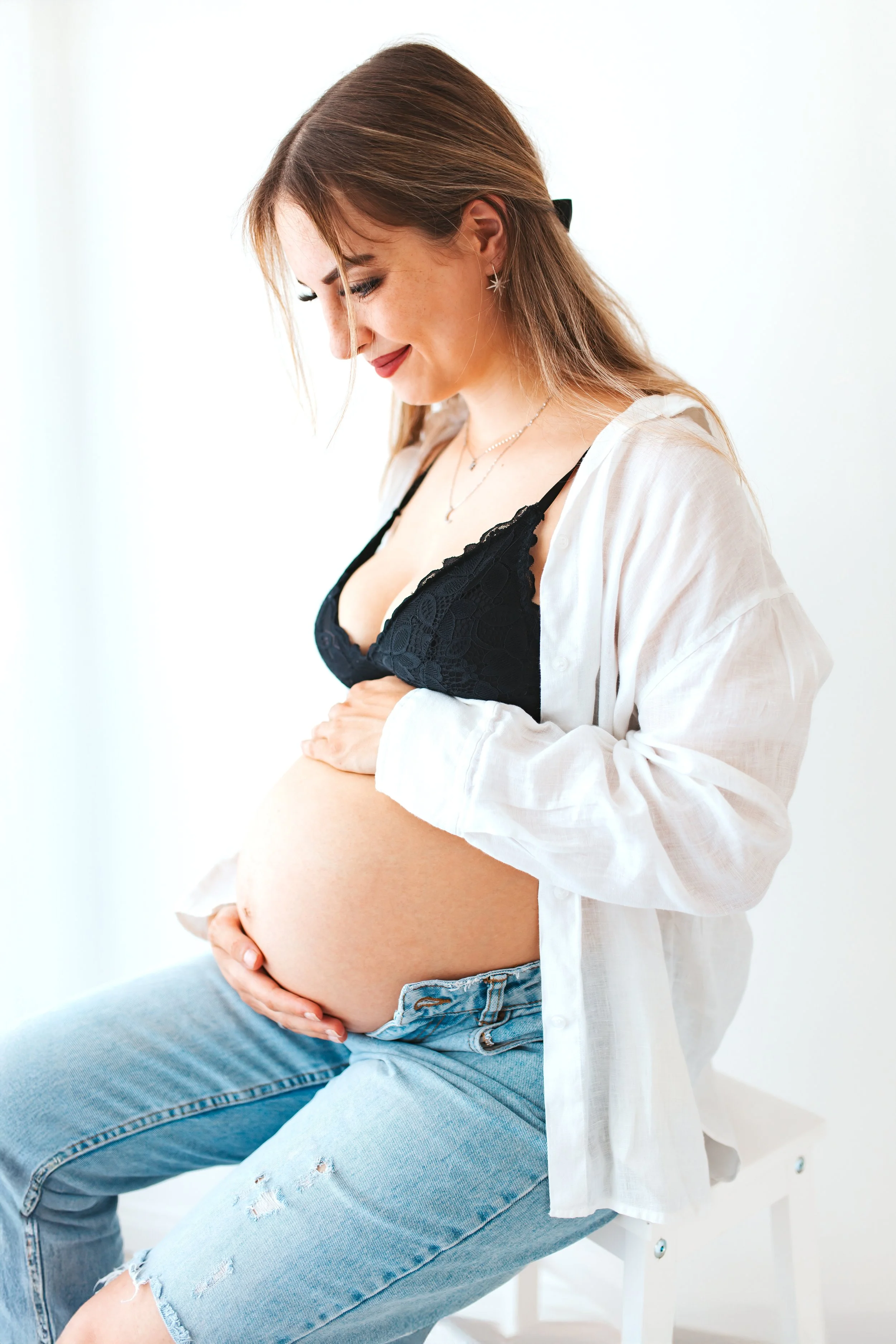 Schwangere Frau sitzt auf einem weißen Stuhl, trägt eine schwarze Spitze-Bademantel und zerrissene Jeans, hält ihren Babybauch mit beiden Händen, lächelt und schaut nach unten.