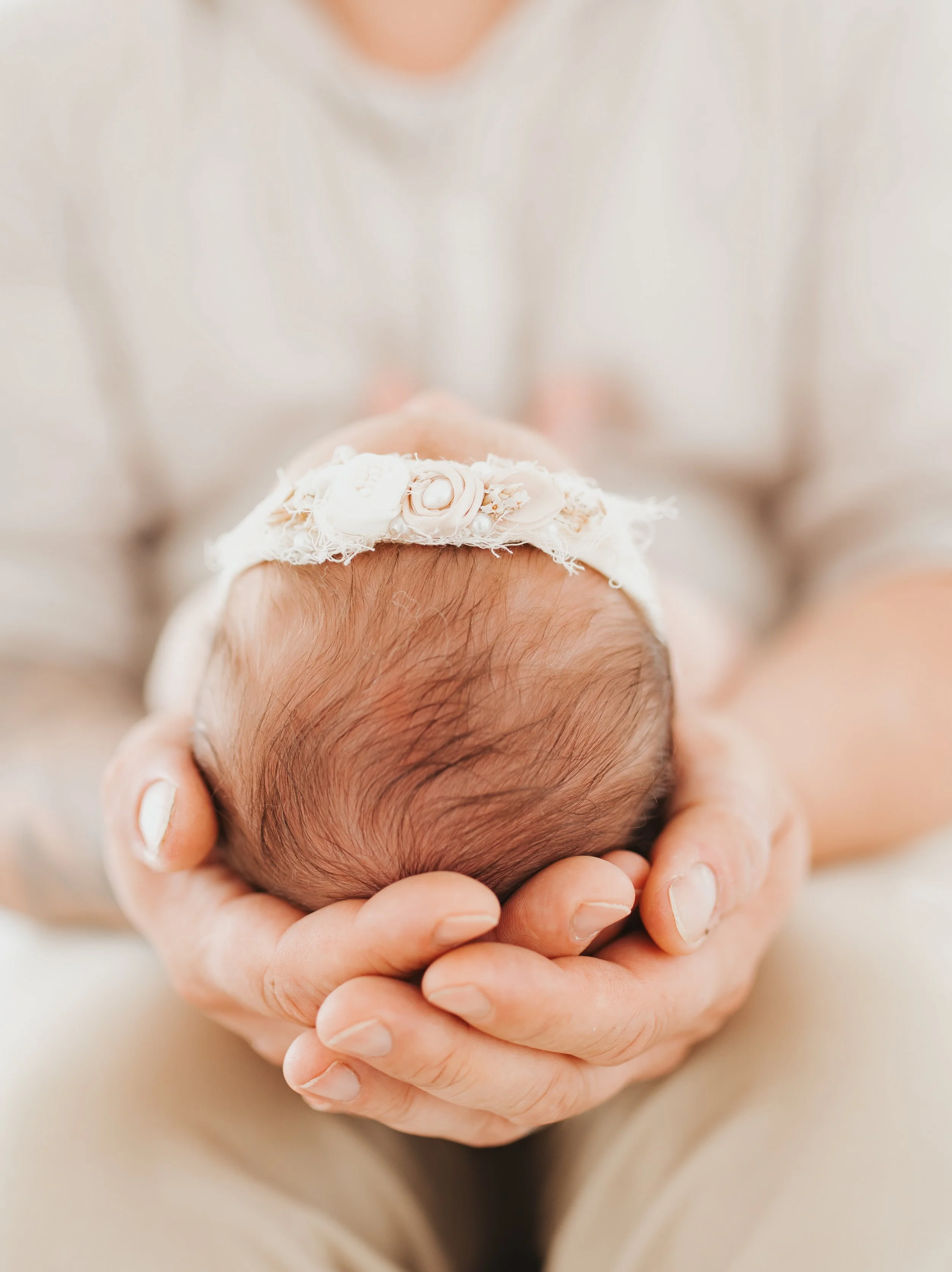Eine Person hält ein neugeborenes Baby mit einem weißen Kopfschmuck in den Händen. Das Baby liegt auf den Händen, der Fokus liegt auf dem Kopf des Babys.
