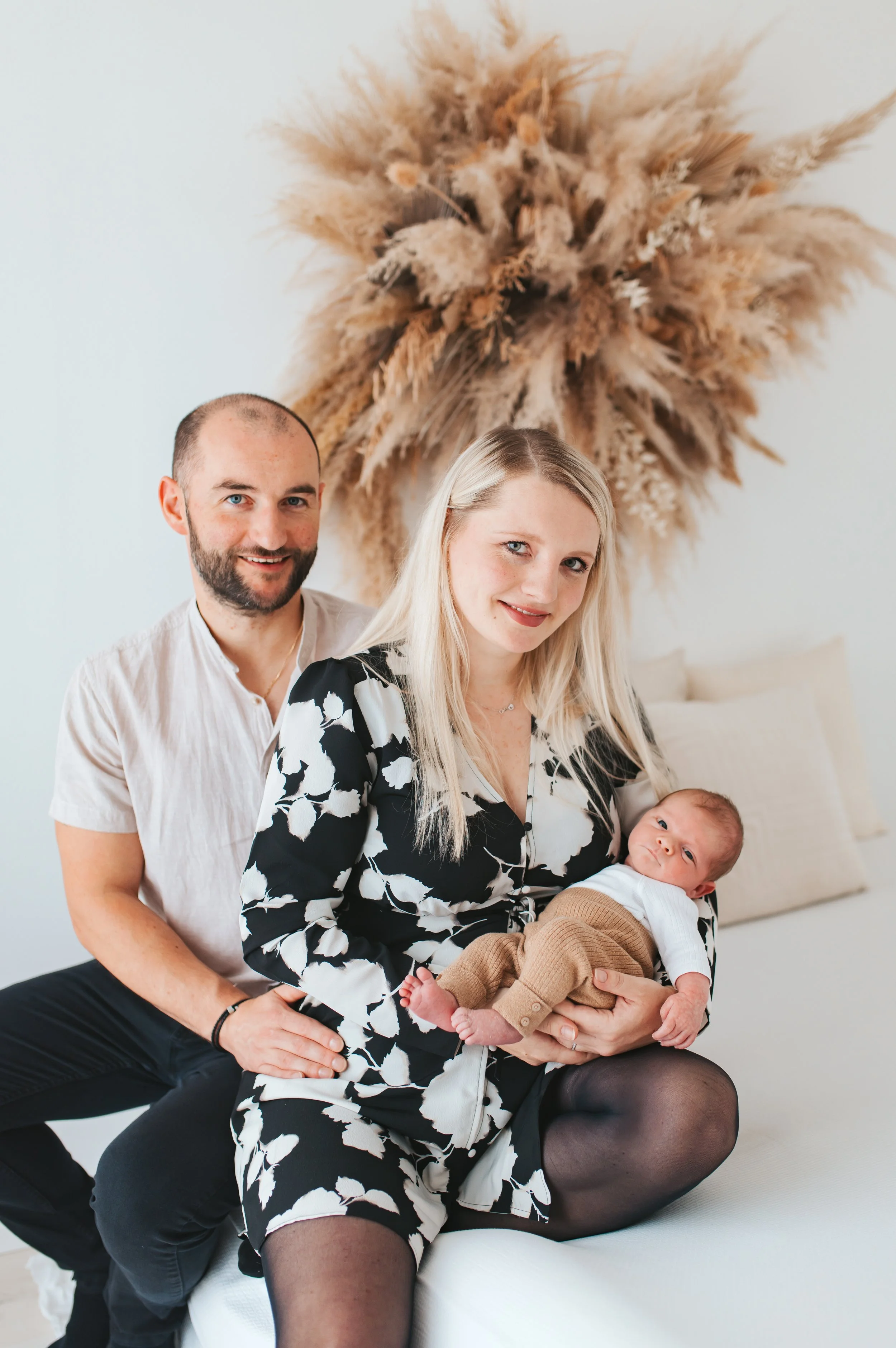 Eine junge Familie mit einem Baby sitzt auf einem Bett. Ein Mann, eine Frau und ein Neugeborenes. Im Hintergrund eine dekorative Wand mit Trockenzweigen.