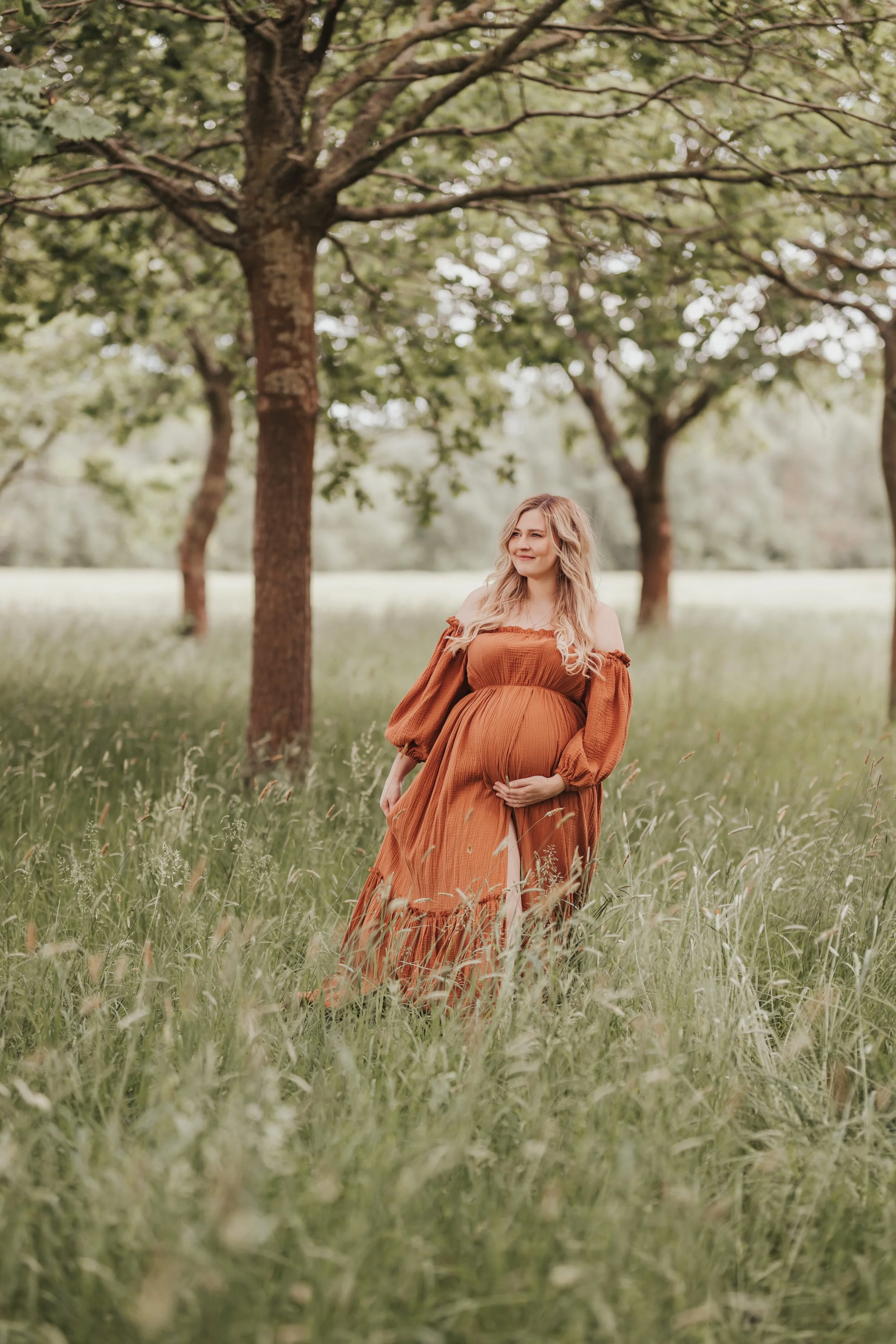 Schwangere Frau in einem orangefarbenen Kleid steht in einem grünen Wiesenbereich unter Bäumen im Freien.