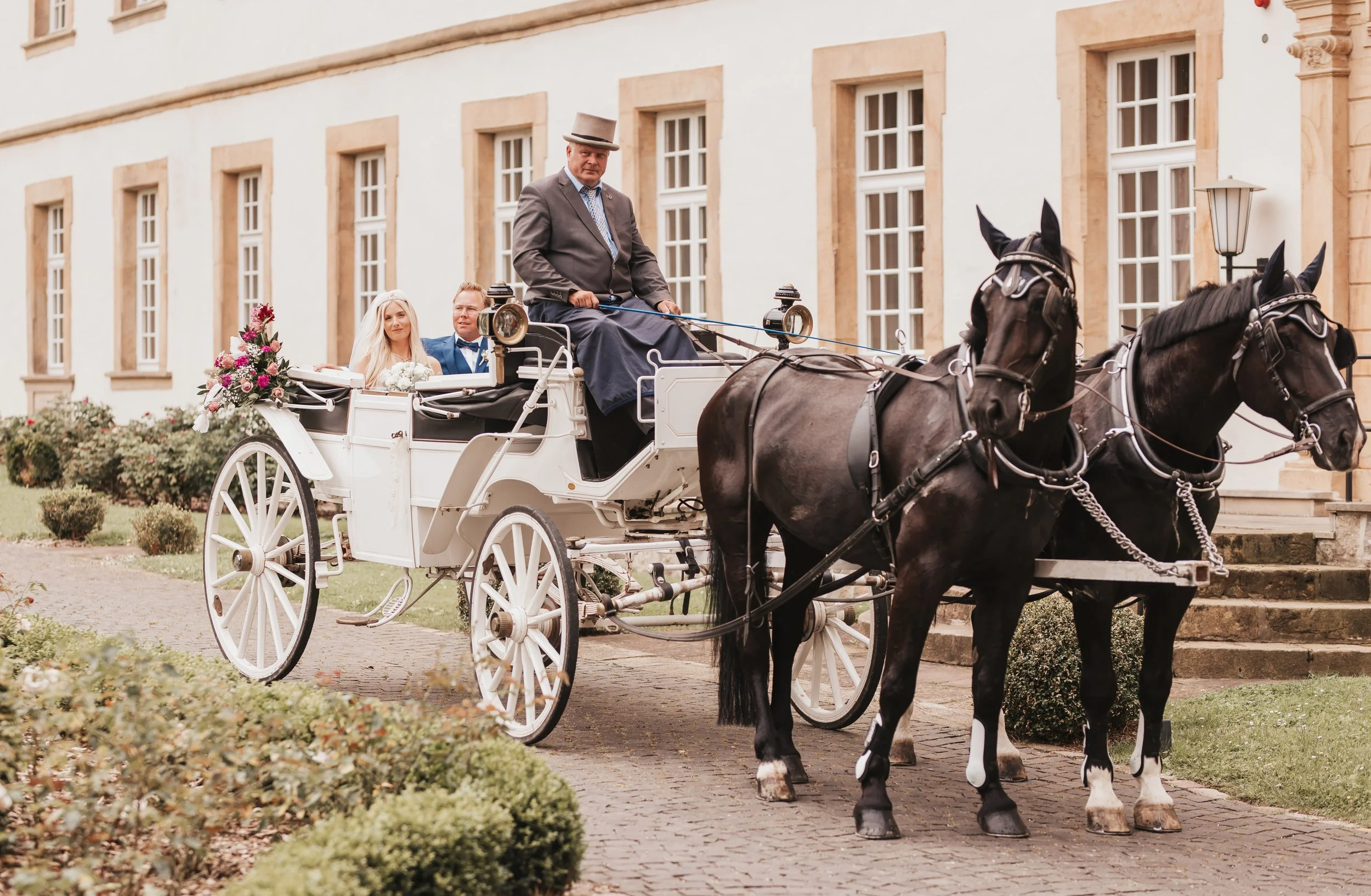 Brautpaar in einem weißen Kutschwagen vor einem historischen Gebäude, gezogen von zwei schwarzen Pferden, bei einer Trauung im Freien.