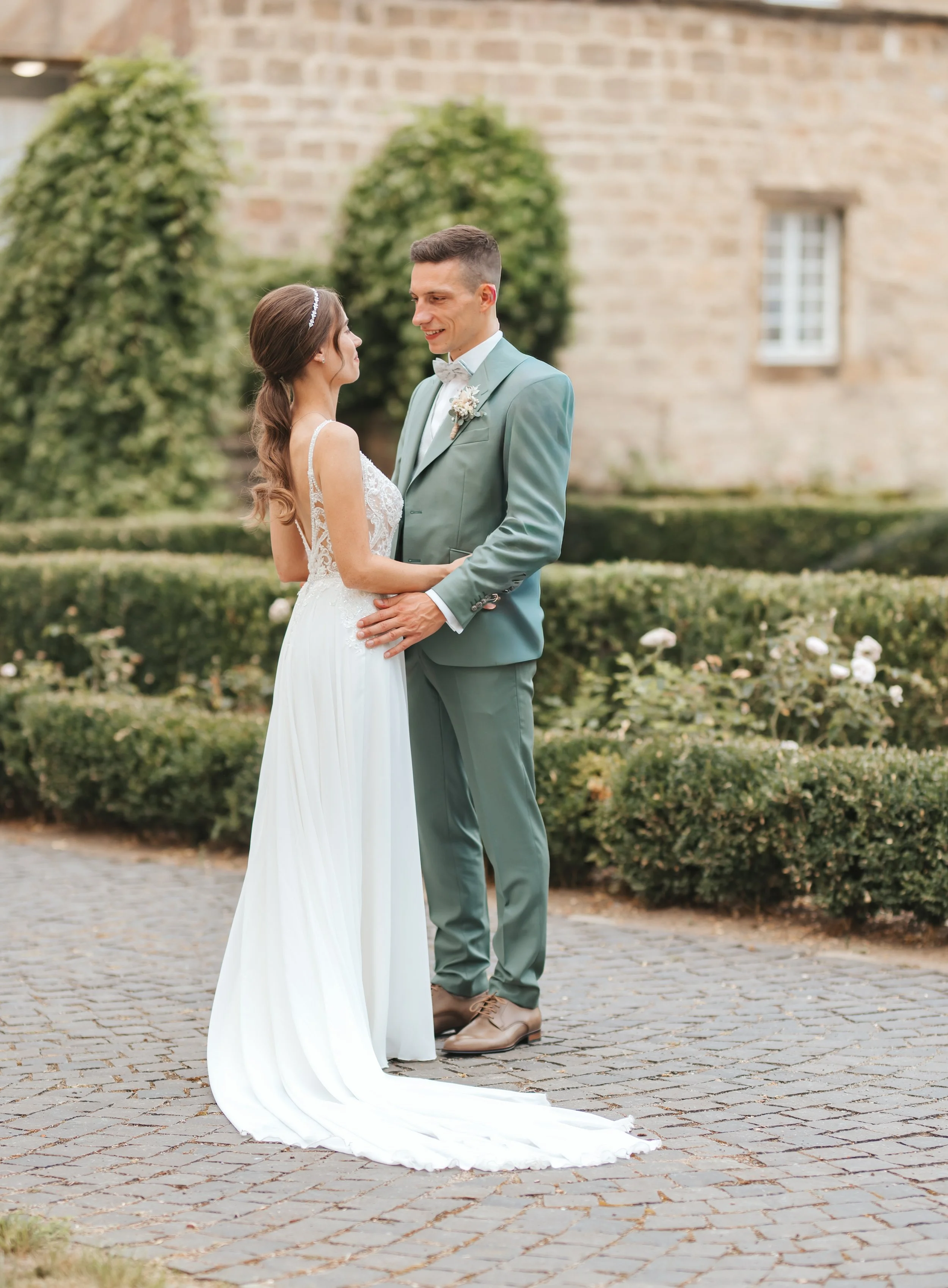 Ein Brautpaar steht sich bei ihrer Hochzeit im Garten gegenüber, die Frau trägt ein weißes Brautkleid mit Schleppe und der Mann einen hellblauen Anzug, umgeben von Blüten und Gartenmotiven.