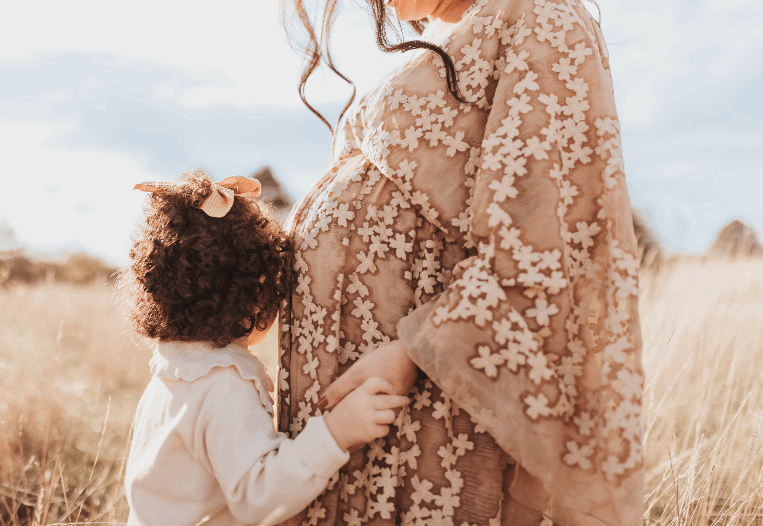 Eine schwangere Frau hält die Hand eines jungen Mädchens in einem Feld, beide tragen beige Kleidung und die Frau trägt ein Kleid mit Blumenmuster.