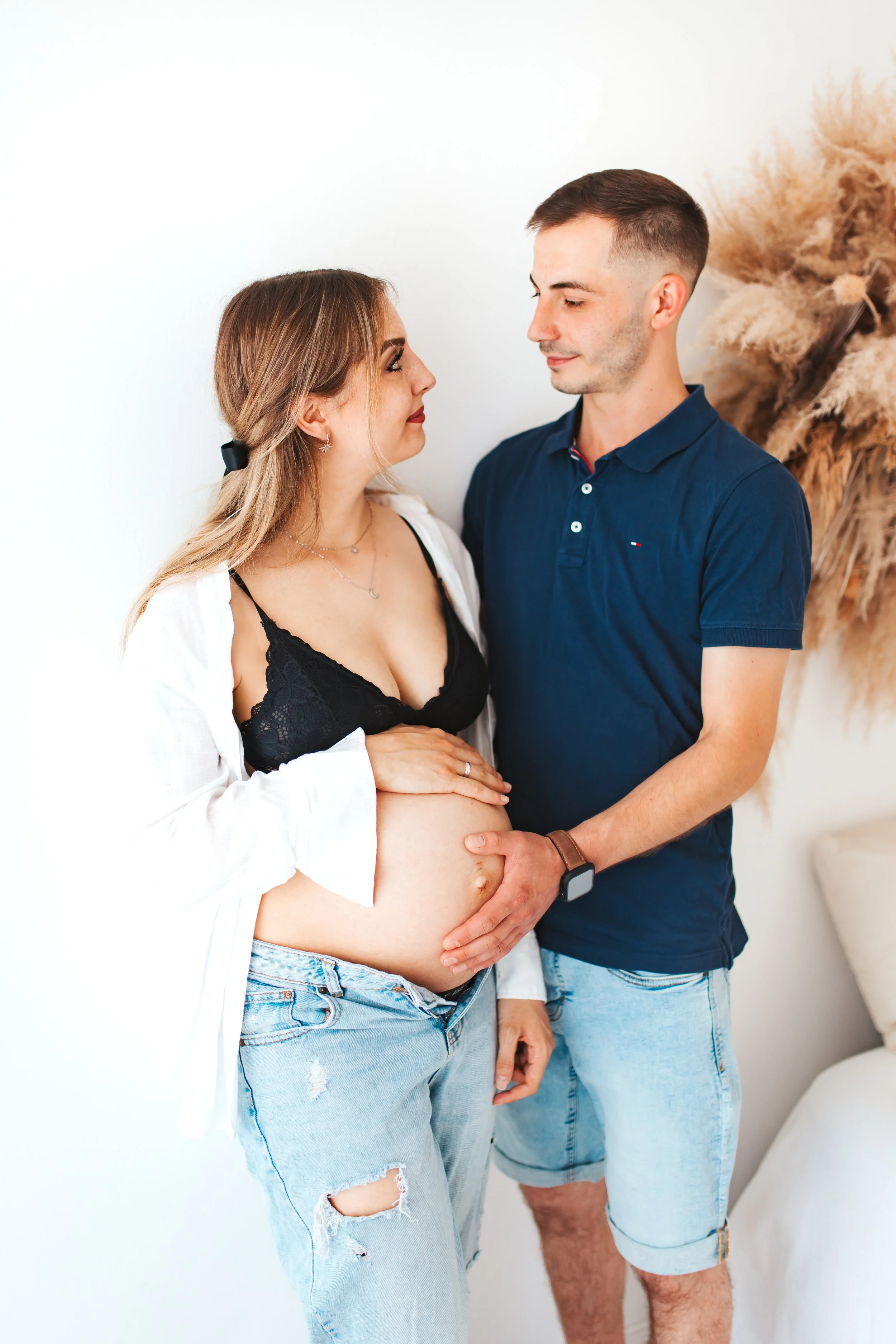 Ein schwangertes Paar schaut sich liebevoll an, während der Mann die Babybauchfrau anlächelt, im Wohnzimmer vor einem weißen Wandhintergrund, mit getrockneten Pflanzen im Hintergrund.