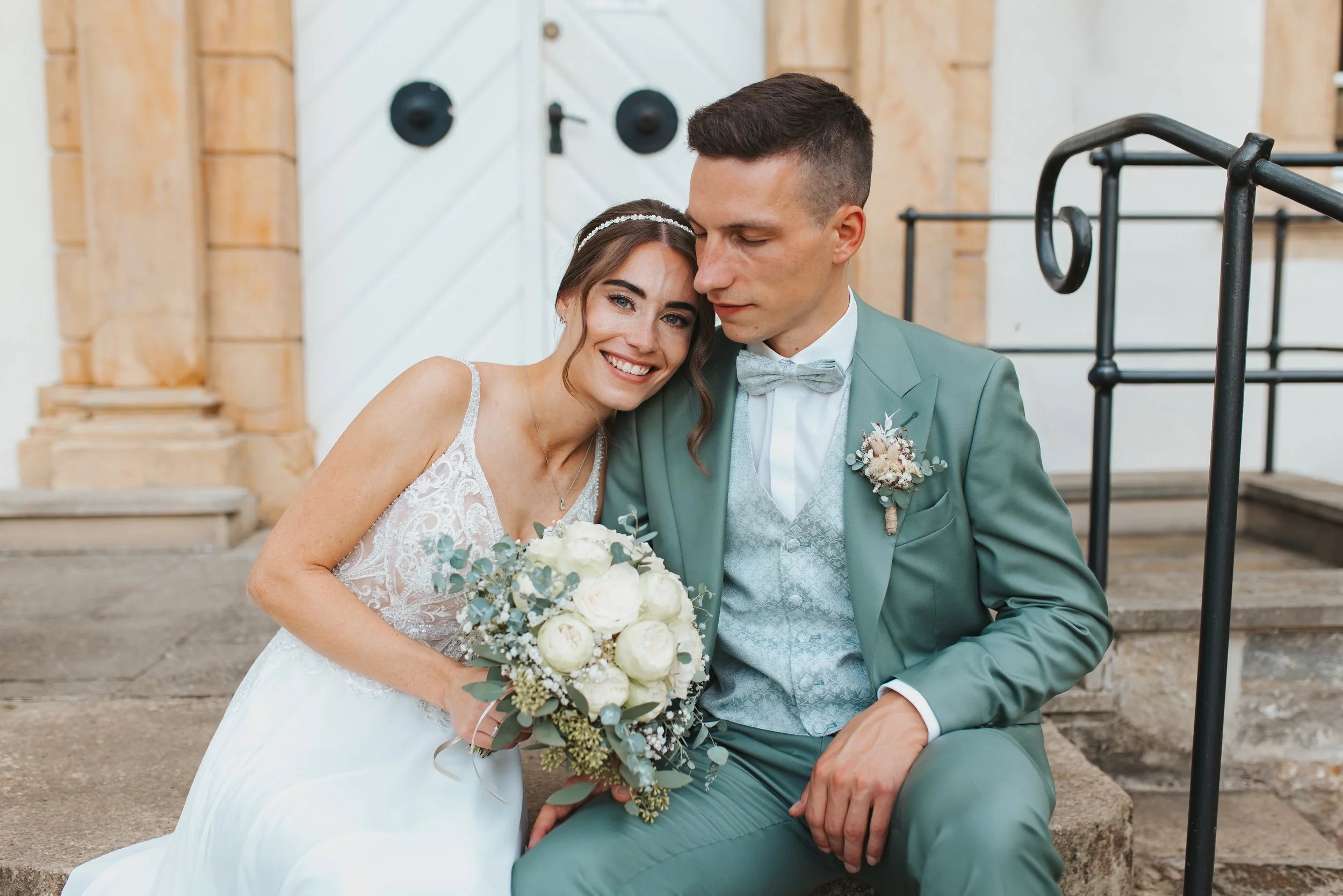 Ein Brautpaar, das auf den Stufen eines alten Gebäudes sitzt. Die Braut trägt ein weißes Hochzeitskleid und hält einen Brautstrauß, während der Bräutigam in einem grünen Anzug sitzt. Beide erscheinen glücklich und innig, die Braut lächelt.