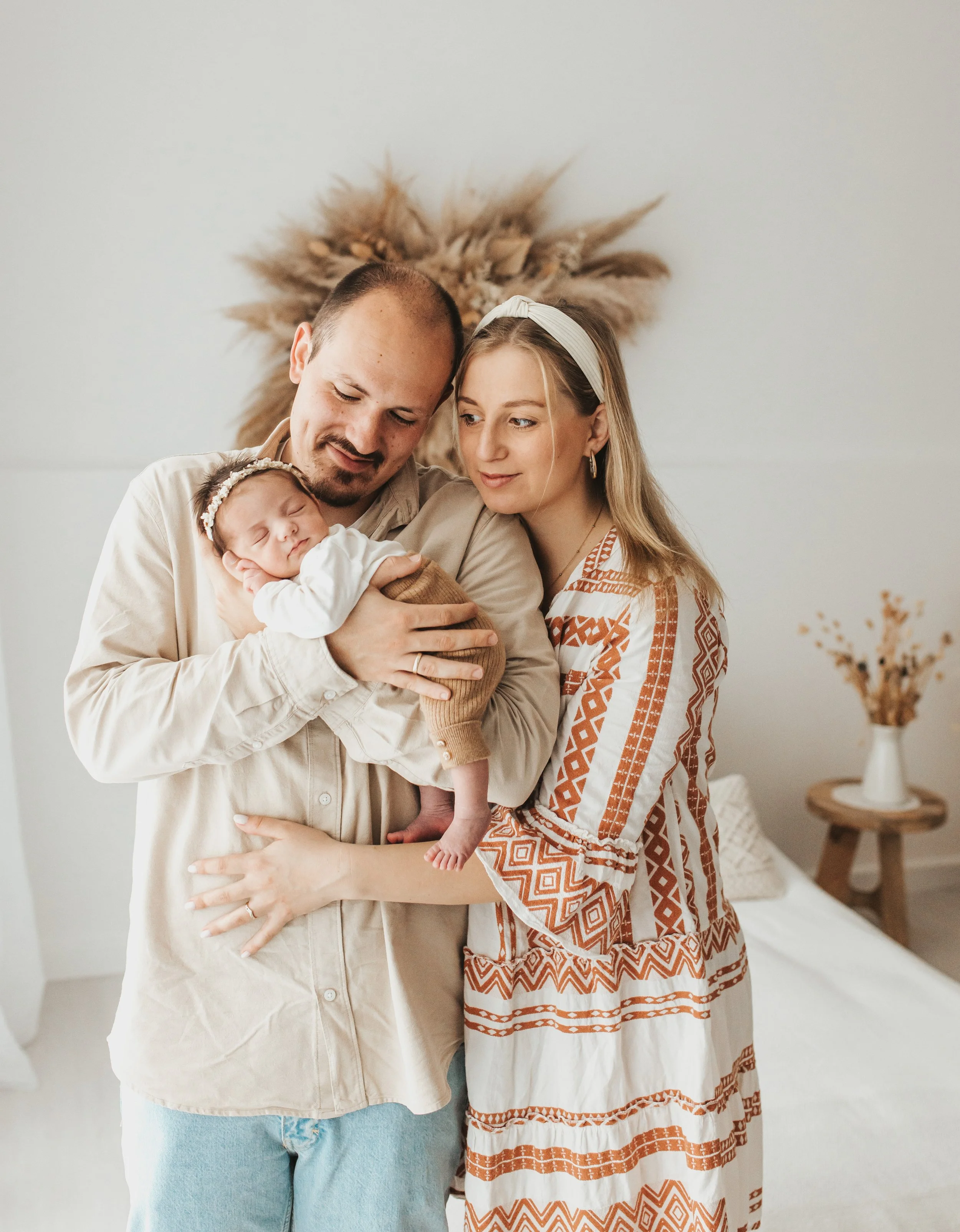 Eine junge Familie mit einem Neugeborenen, die im Schlafzimmer lächelnd zusammenstehen.