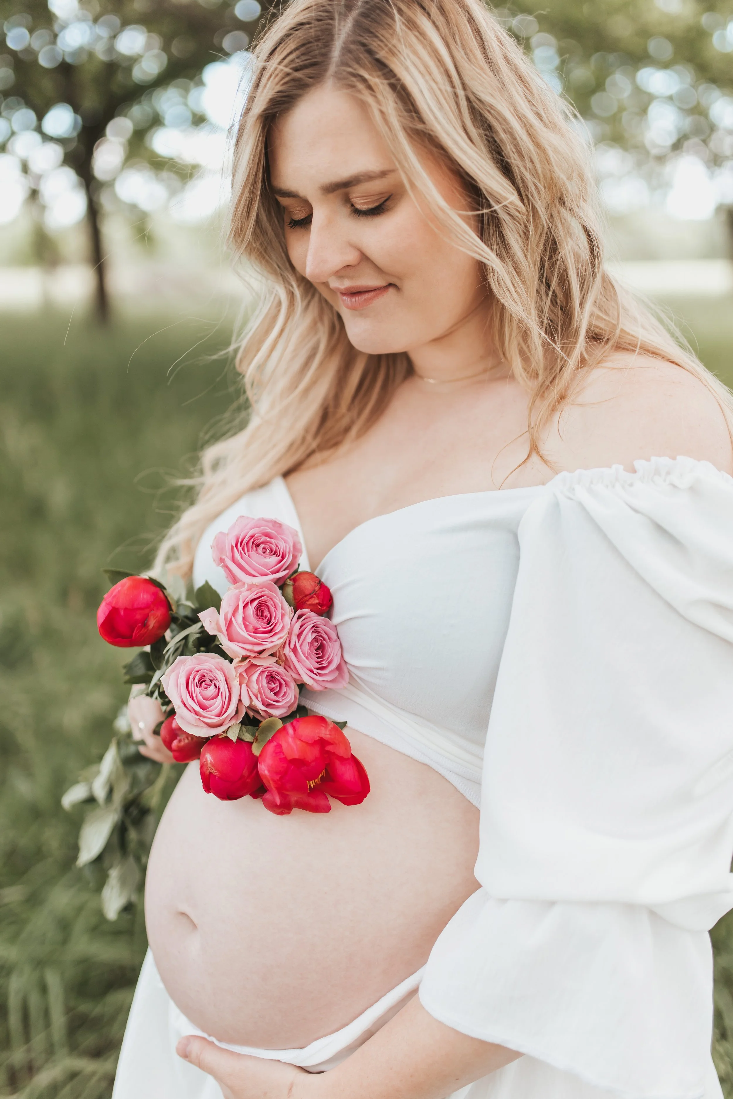 Schwangere Frau mit Blumen auf dem Bauch im Freien, lächelnd mit geschlossenem Blick.
