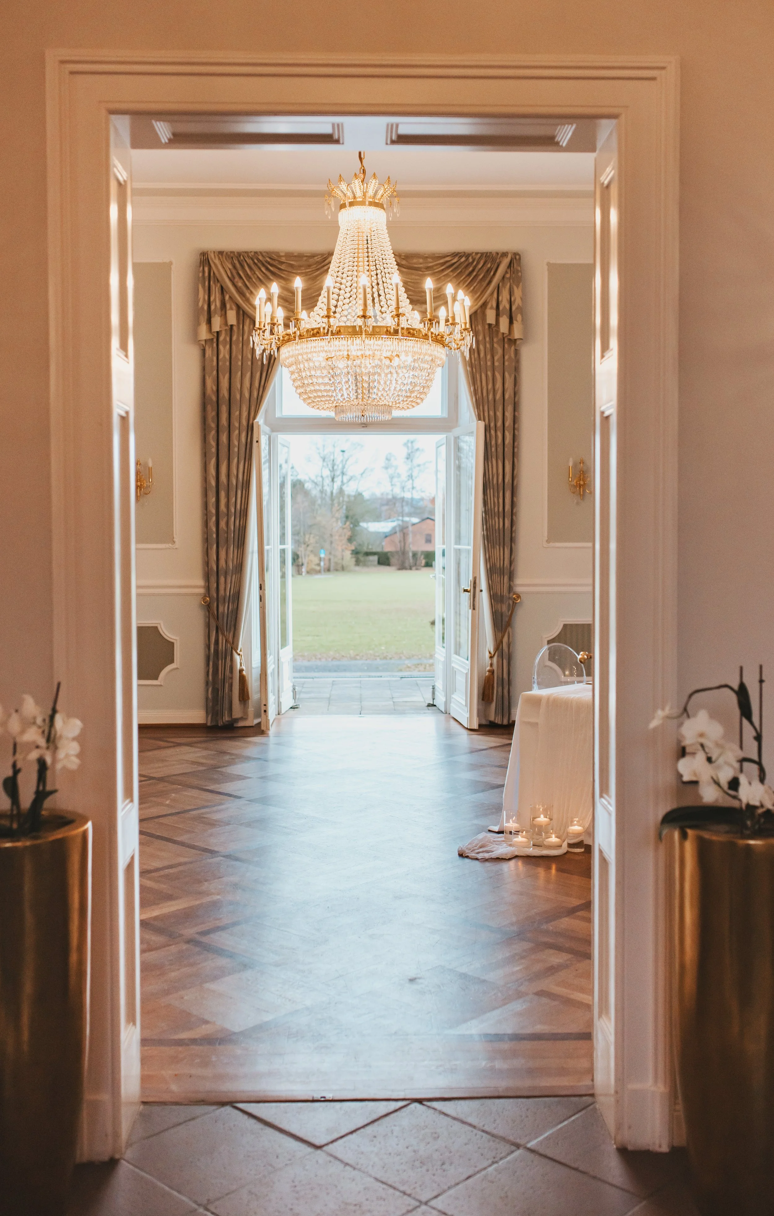 Ein eleganter Raum mit einem großen Kronleuchter, französischen Türen, die nach draußen auf ein grünes Gelände öffnen, und einem großen Fenster mit goldfarbenen Vorhängen.