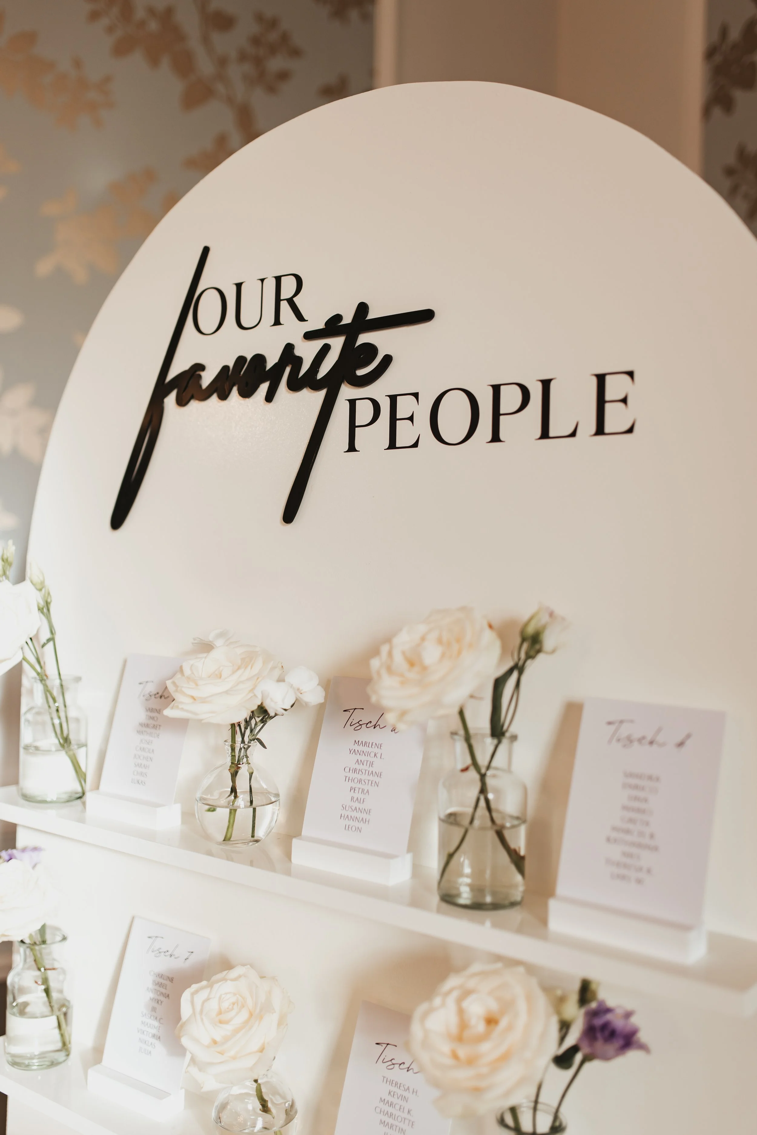 Ein Bücherstapel mit weißen Rosen und Namensschildern auf einer weißen Oberfläche, vor einer großen weißen Wand mit schwarzem Schriftzug 'Our favourite people'.