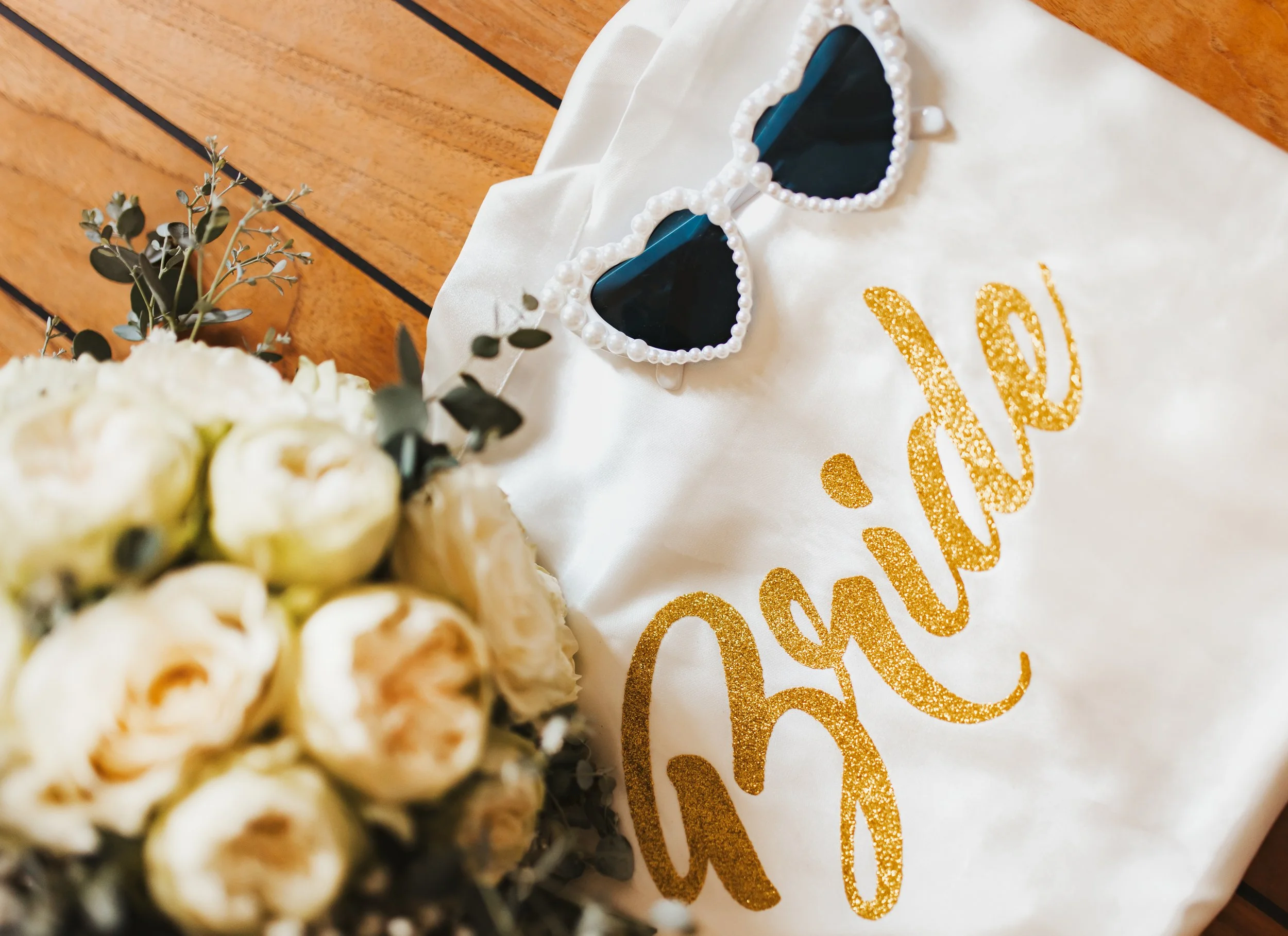 Weißes Tuch mit goldglitternder Aufschrift 'love', Sonnenbrille mit weißem Perlenrahmen, Blumenstrauß mit weißen Rosen und Grün, auf Holz-Tisch