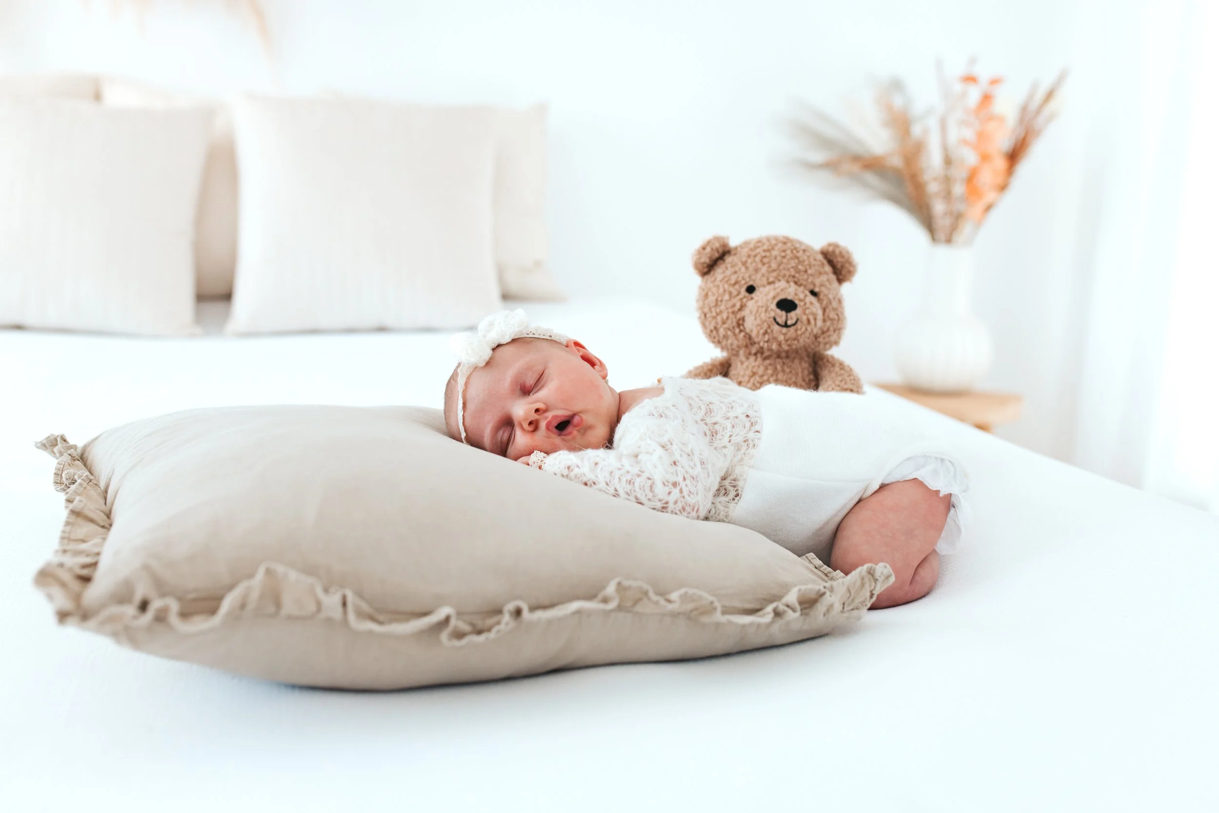 Ein schlafendes Baby liegt auf einem großen Kissen in einem hellen, neutral eingerichteten Raum. Im Hintergrund steht ein niedlicher Teddybär und eine Vase mit Trockenblumen.