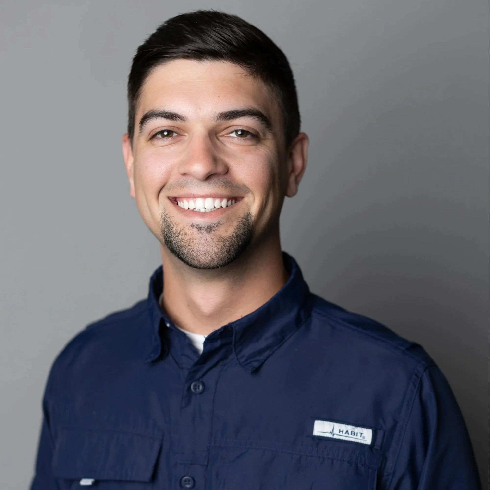 Close-up portrait of a smiling man with dark hair, facial hair, and light skin, wearing a navy blue shirt with a name tag that says "HABIT" on a gray background.