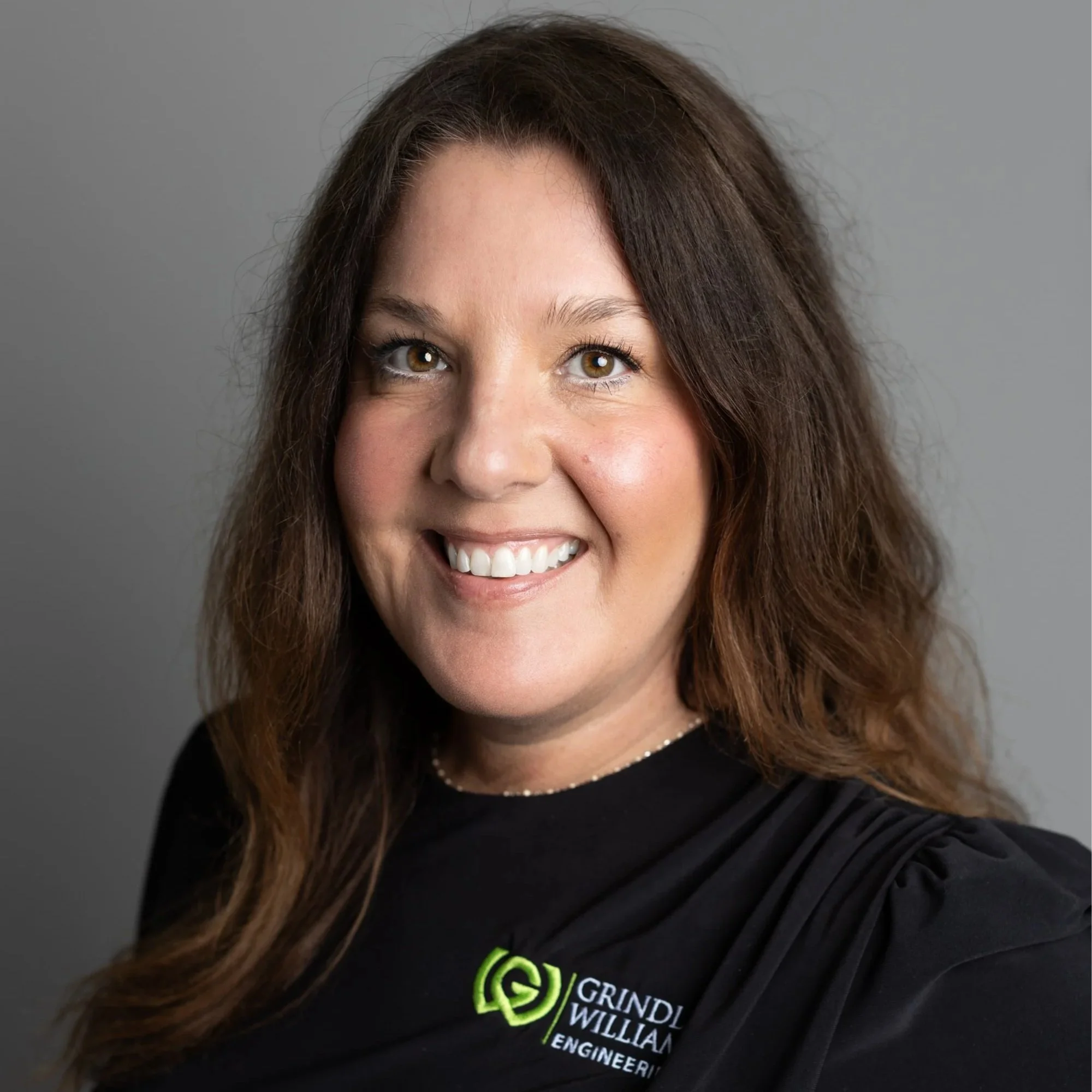 Portrait of a smiling woman with curly red hair, wearing a black sleeveless shirt and sporting tattoos on her arms against a gray background.