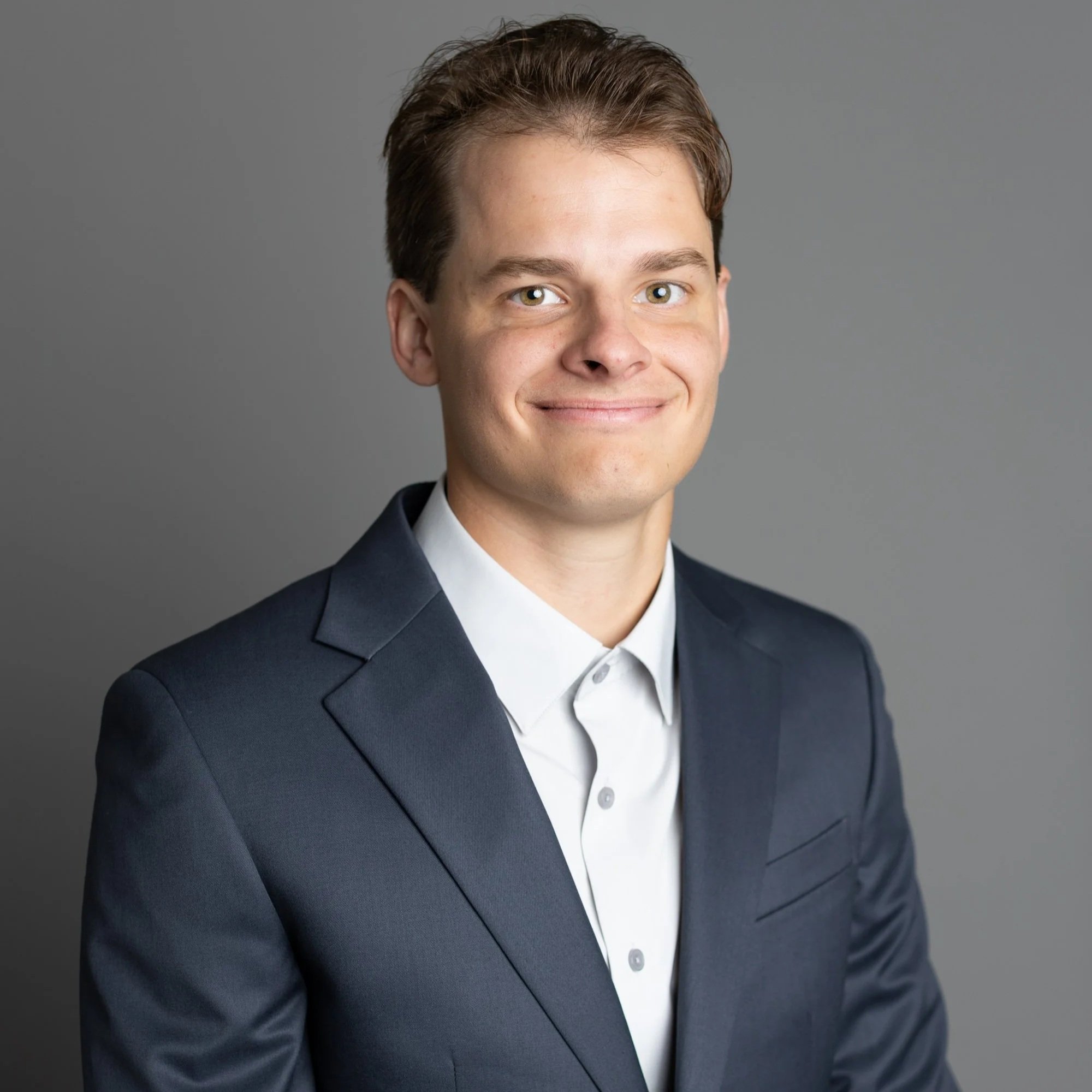 Headshot of a young man with short brown hair, fair skin, and green eyes. He is smiling and dressed in a dark suit and white shirt against a gray background.