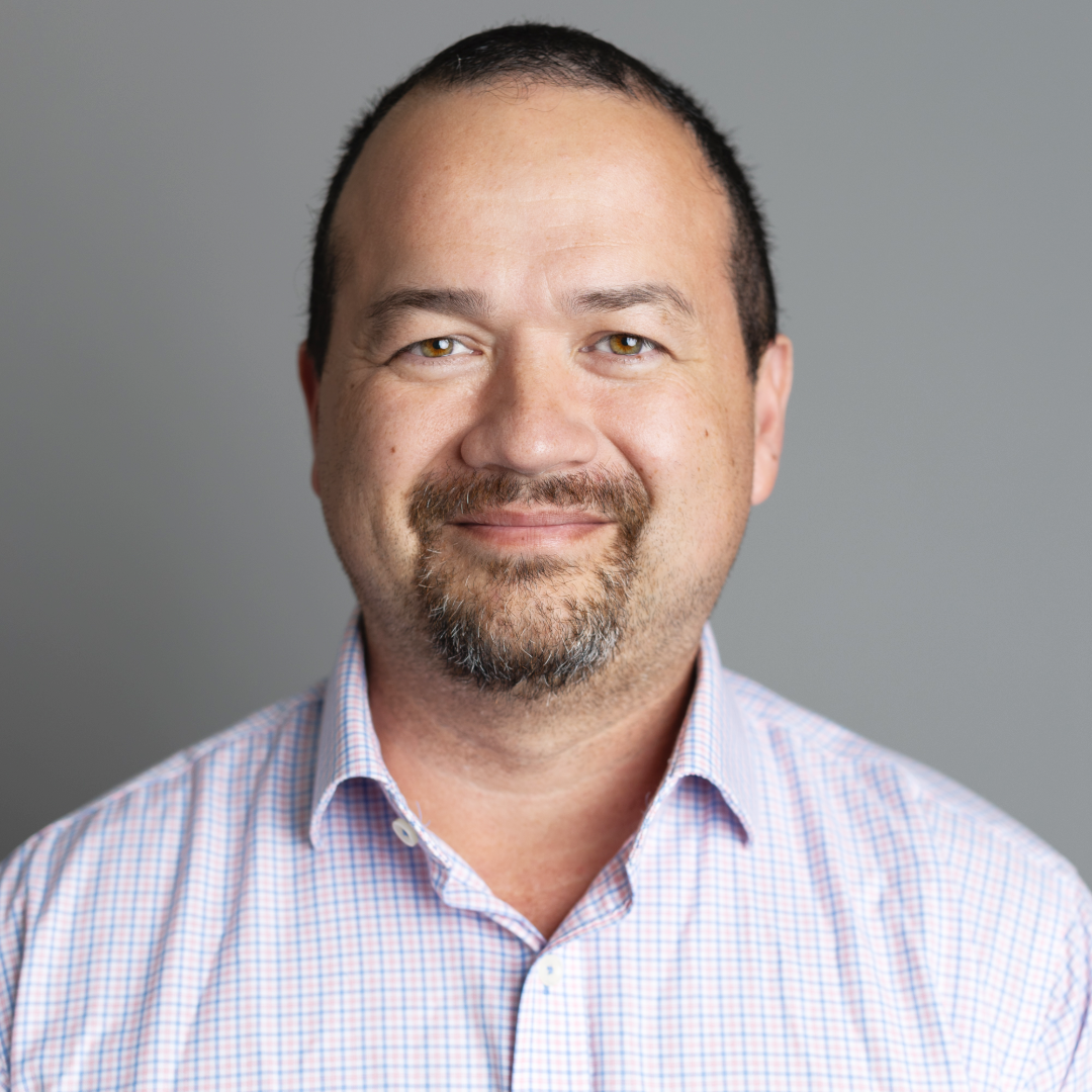 A headshot of a middle-aged man with dark hair, a trimmed beard, wearing a light-colored, checked dress shirt, smiling softly against a neutral gray background.