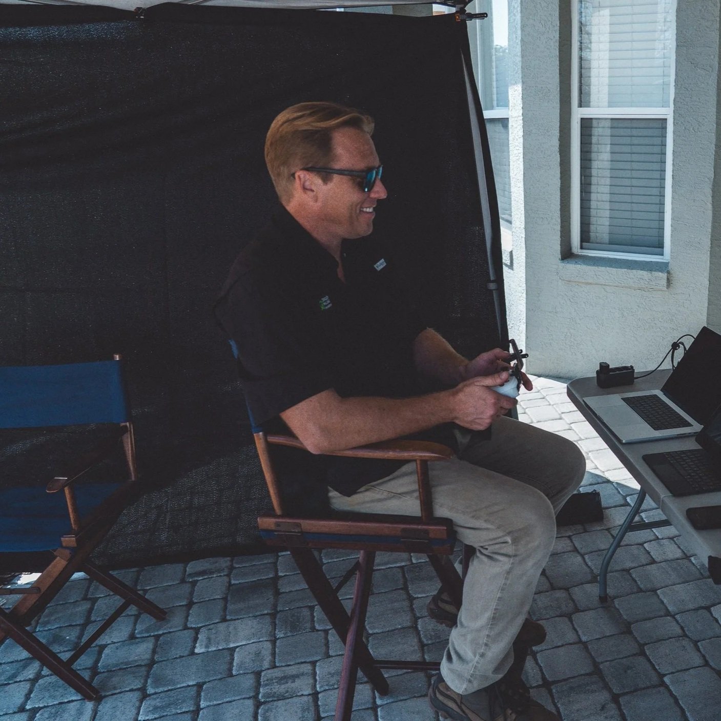 A man sitting outdoors on a patio, wearing sunglasses and smiling while using a drone remote controller, with a laptop and other electronic devices on a table nearby.