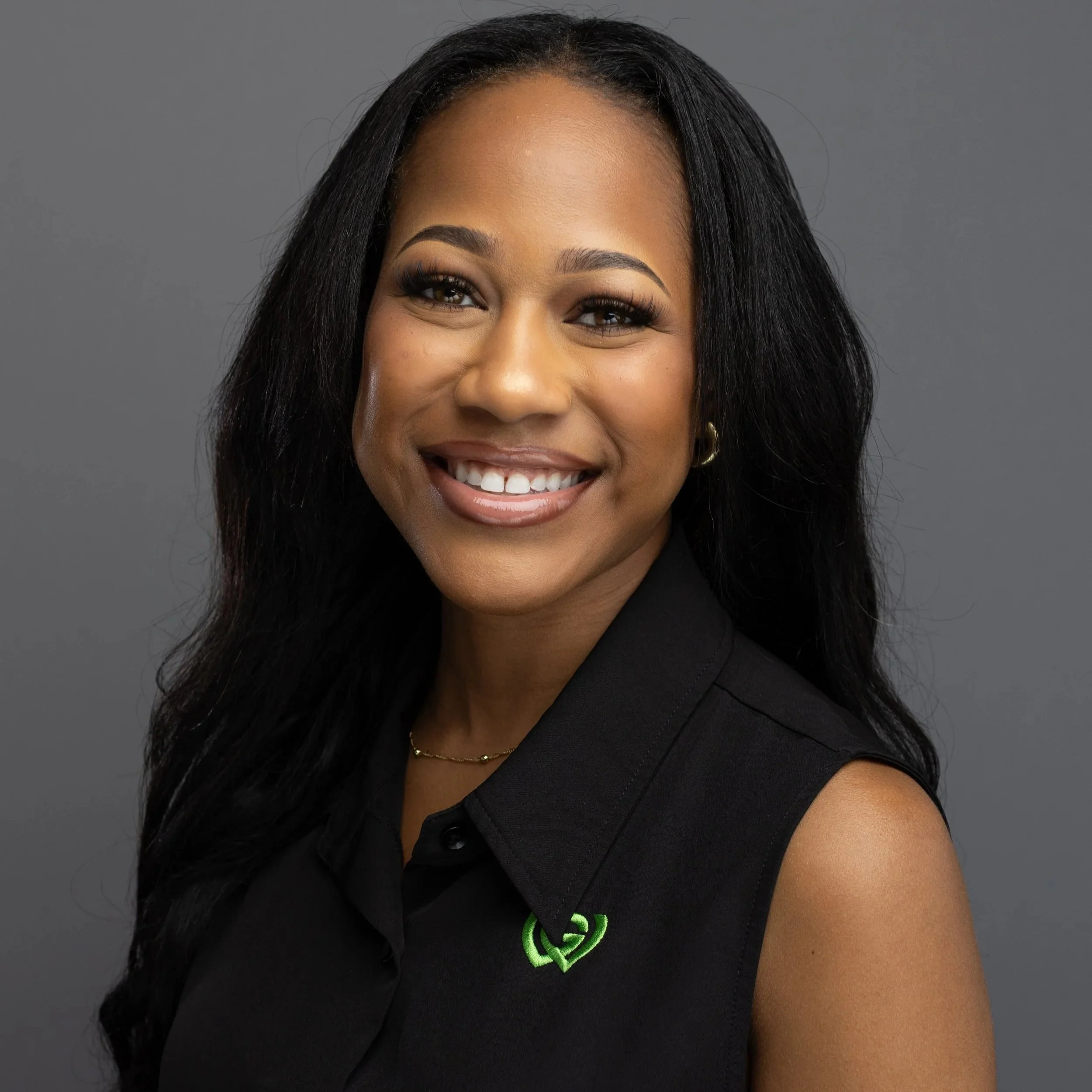 A smiling young African American woman with long black hair wearing a black sleeveless shirt and small hoop earrings, standing against a plain dark gray background.