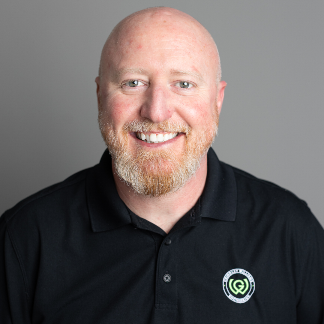 A smiling man with a red beard, bald head, and blue eyes wearing a black polo shirt with a green and white logo on the chest, standing against a gray background.