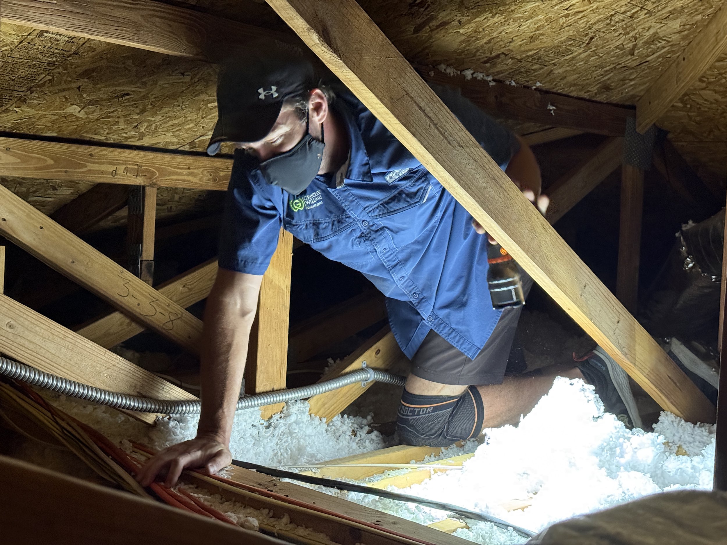 Man wearing a black cap, face mask, and blue work shirt inspecting the wooden framing of an attic or crawl space, with insulation and electrical wiring visible.